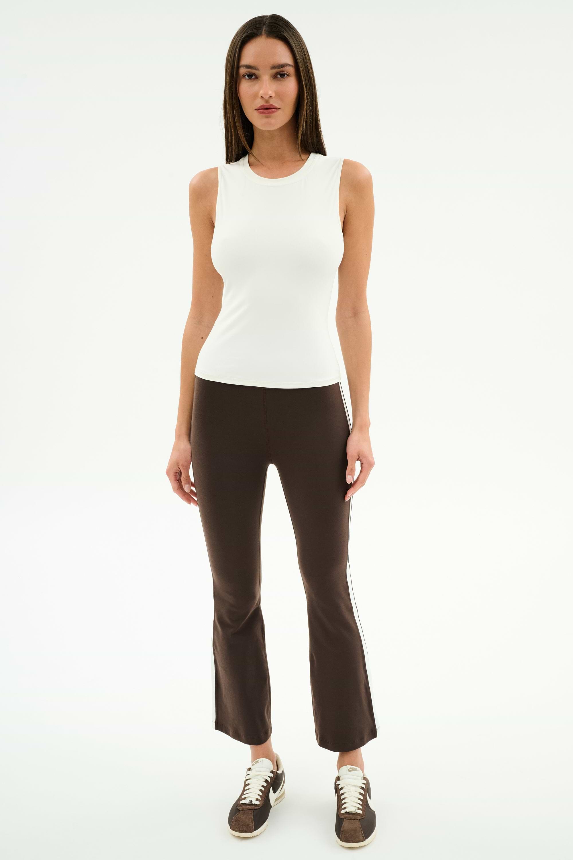 A woman faces forward in the SPLITS59 Raquel High Waist Supplex Crop in Dark Chocolate/White, styled with a sleeveless white top and brown sneakers, standing against a plain white background.