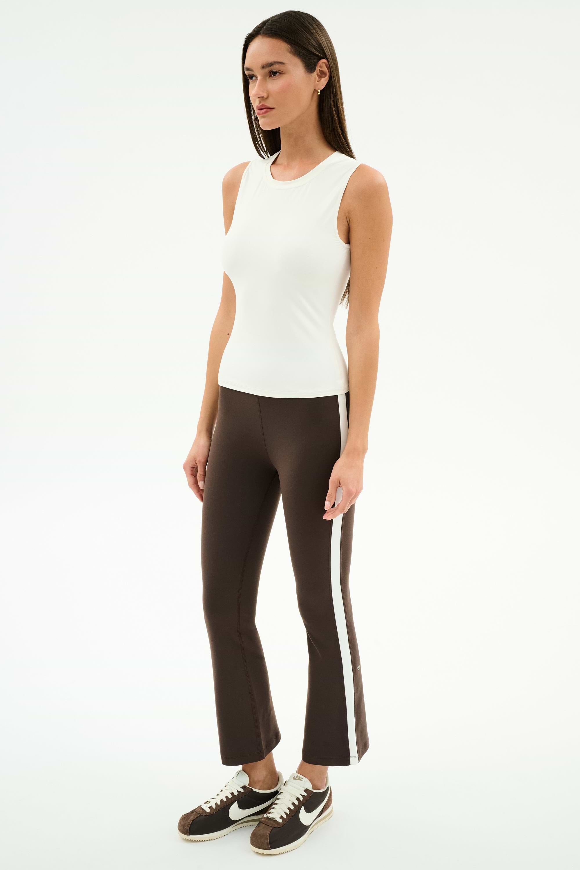A woman stands against a plain white background wearing the SPLITS59 Raquel High Waist Supplex Crop in Dark Chocolate/White and a white sleeveless top, paired with brown Nike sneakers.