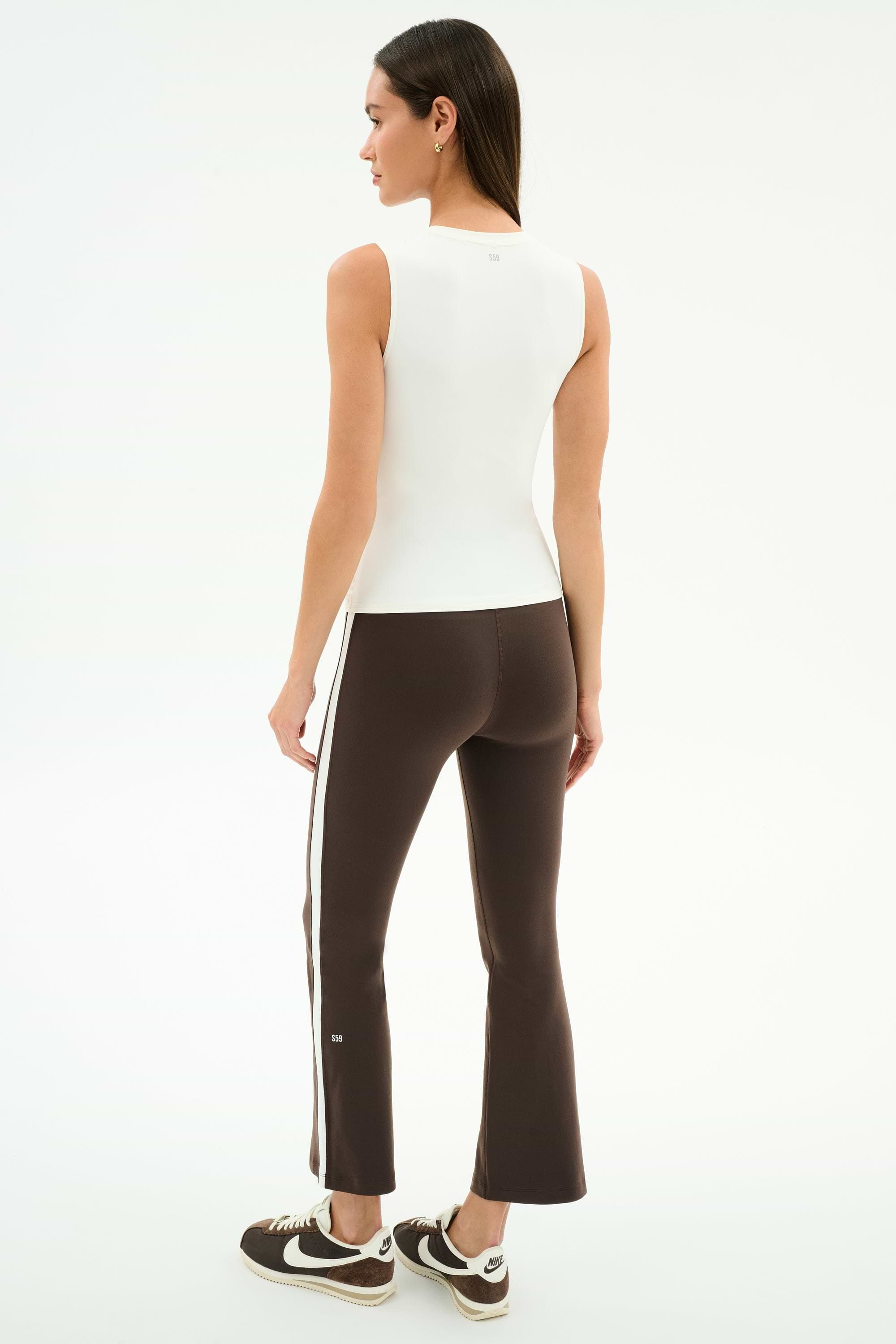 A woman stands facing away, wearing the SPLITS59 Raquel High Waist Supplex Crop in Dark Chocolate/White, a sleeveless white nylon top, and black sneakers against a white background.