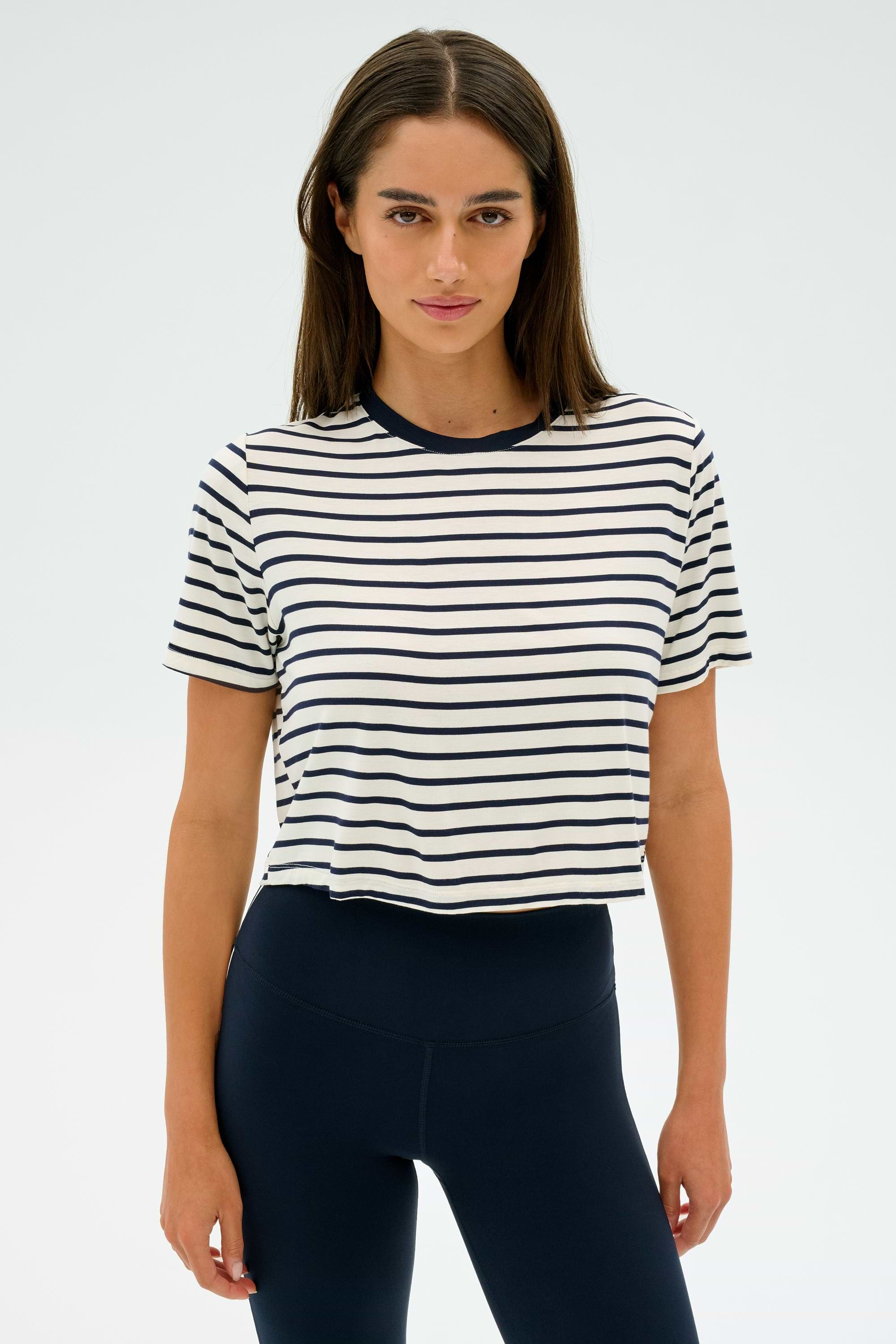 A person wearing the SPLITS59 Djuna Crop Ringer in Indigo Stripe and dark leggings stands against a plain background, ready for gym workouts.