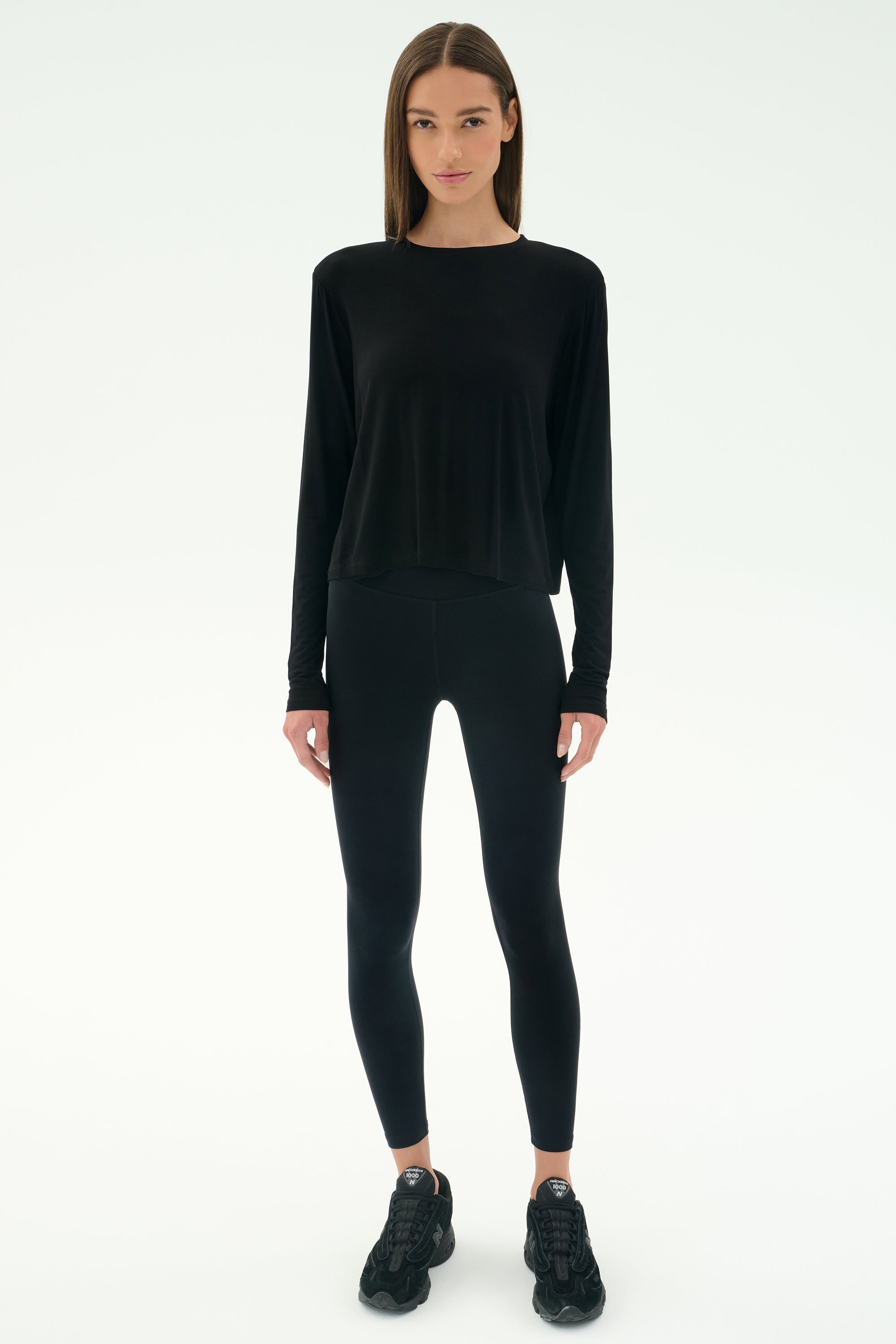 A person stands facing forward in SPLITS59's Djuna Long Sleeve - Black, paired with black leggings and sneakers—ideal activewear for cool weather workouts—against a plain white background.