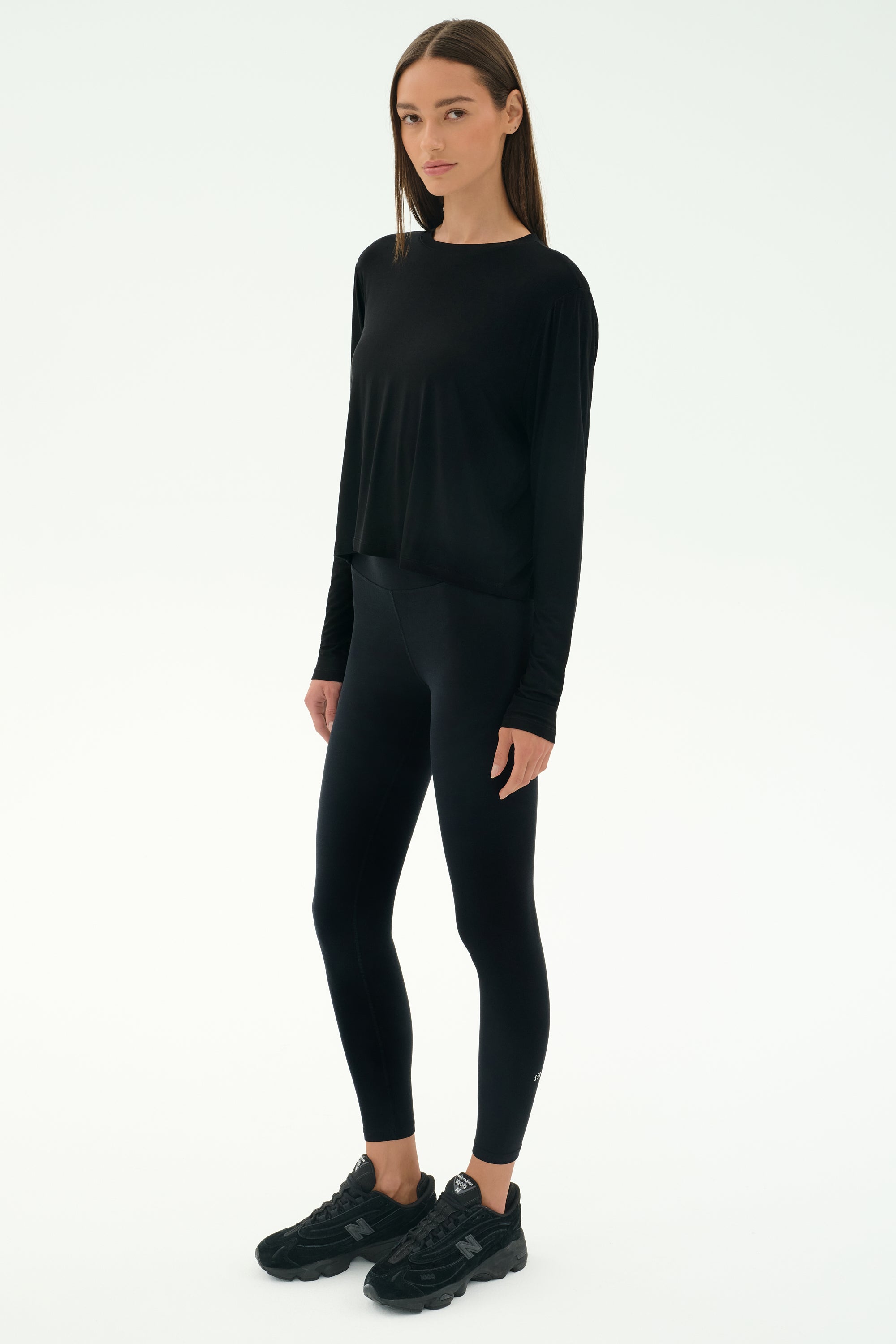 A woman faces forward against a white background wearing the SPLITS59 Djuna Long Sleeve - Black, paired with black leggings and sneakers—an ideal women's activewear look for cool weather workouts.