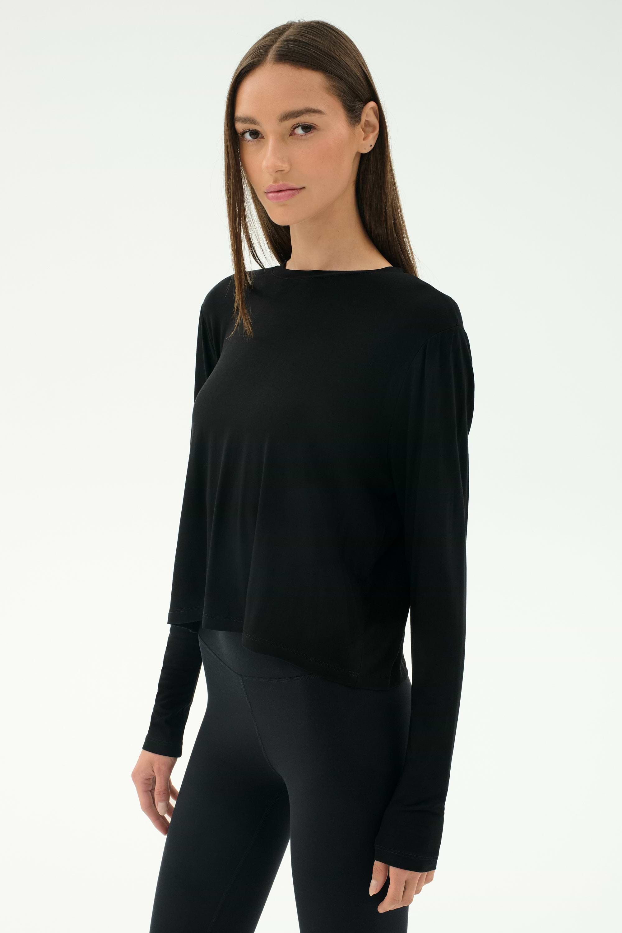 A woman with straight brown hair wears the SPLITS59 Djuna Long Sleeve - Black, paired with black leggings, against a plain white background—ideal for cool weather workouts and stylish women's activewear.