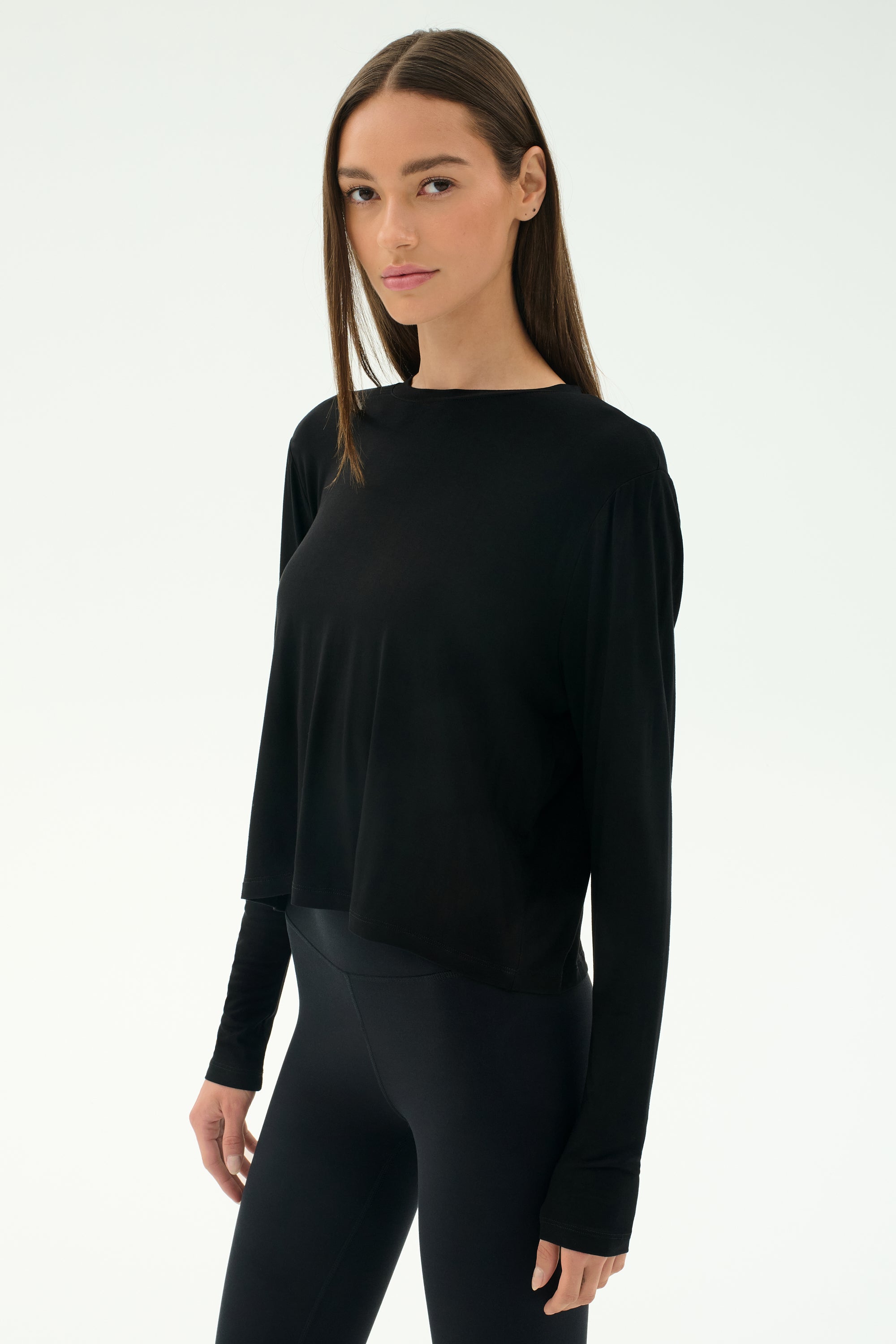 A woman with straight brown hair wears the SPLITS59 Djuna Long Sleeve - Black, paired with black leggings, against a plain white background—ideal for cool weather workouts and stylish women's activewear.