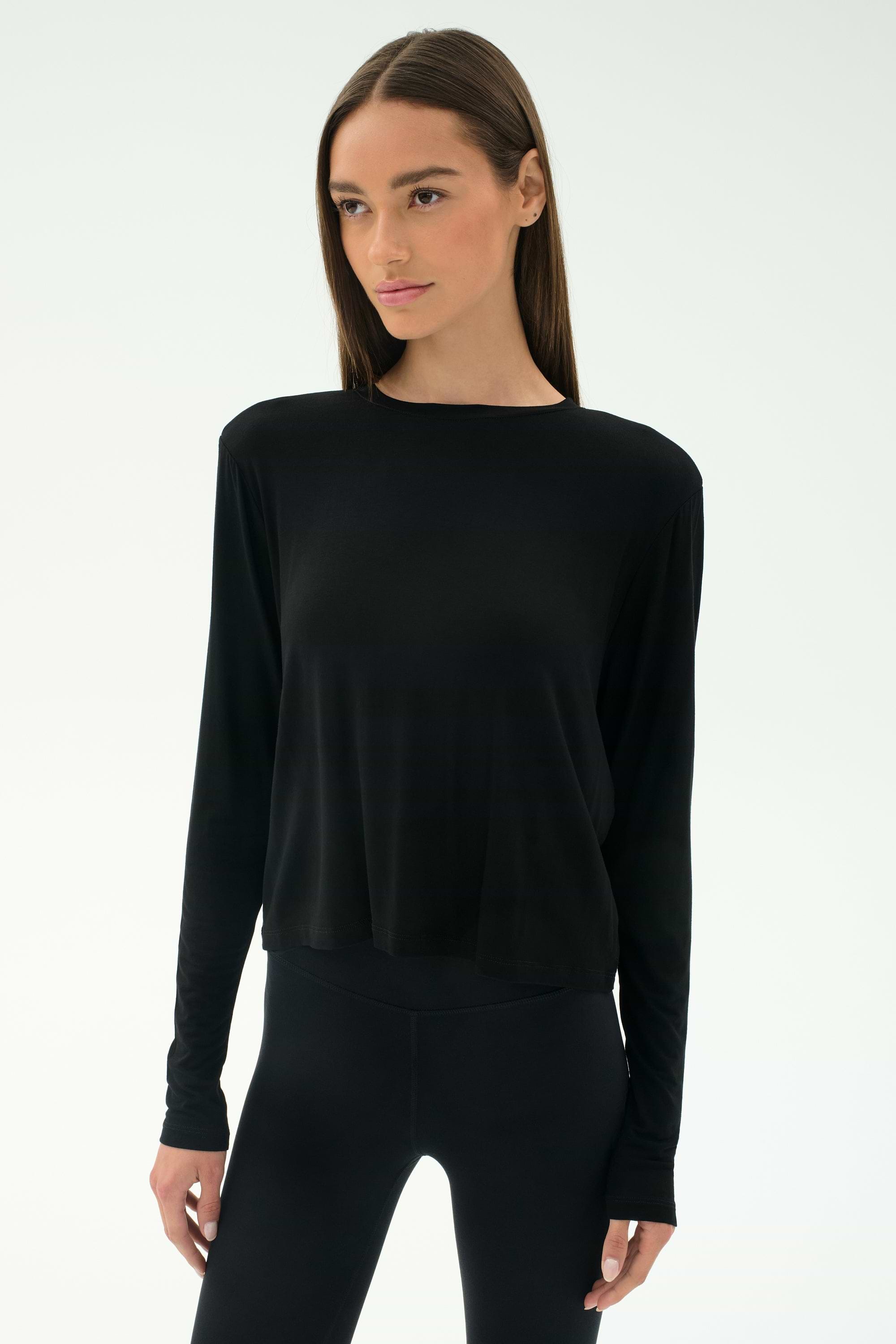 A woman wears the SPLITS59 Djuna Long Sleeve in black, paired with black leggings and standing against a plain light background—ideal women's activewear for cool weather workouts.