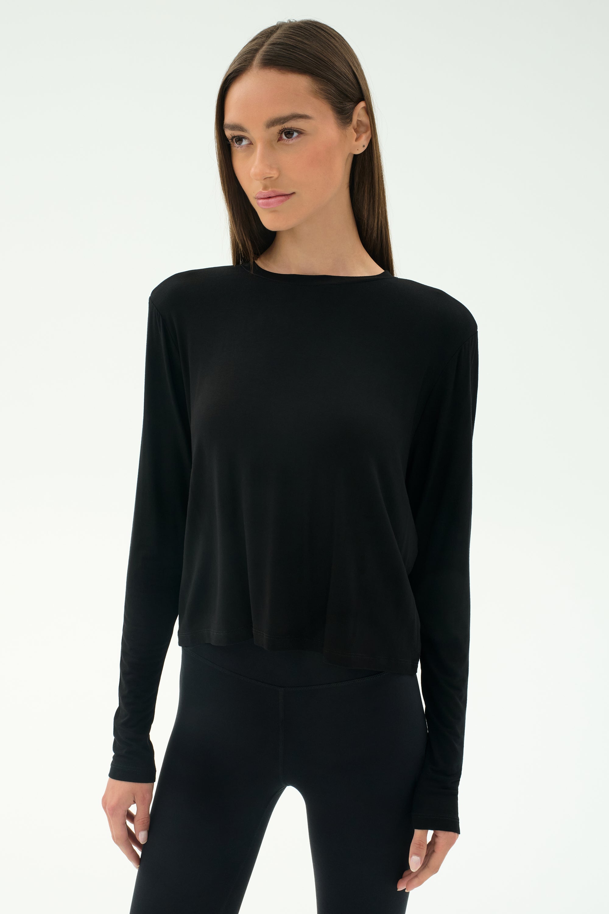 A woman wears the SPLITS59 Djuna Long Sleeve in black, paired with black leggings and standing against a plain light background—ideal women's activewear for cool weather workouts.