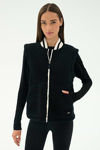 A woman stands against a plain light background, ready for cool weather workouts in the warm SPLITS59 Aster Sherpa Vest - Black, featuring a white zipper and striped collar over a long-sleeve black top and leggings.