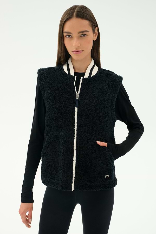 A woman stands against a plain light background, ready for cool weather workouts in the warm SPLITS59 Aster Sherpa Vest - Black, featuring a white zipper and striped collar over a long-sleeve black top and leggings.