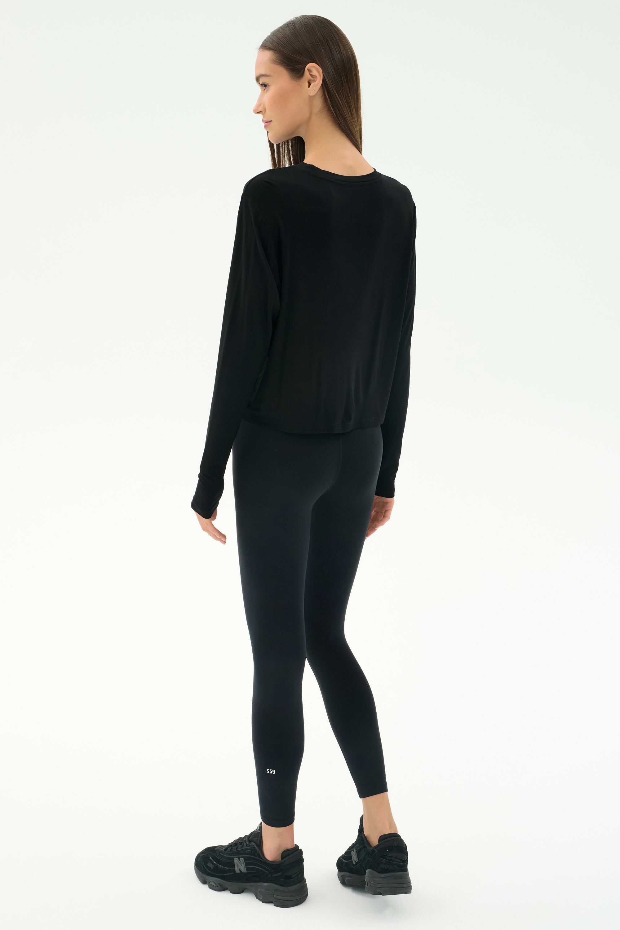 A woman stands on a white background, facing away, wearing the SPLITS59 Djuna Long Sleeve - Black, paired with black leggings and black athletic shoes.