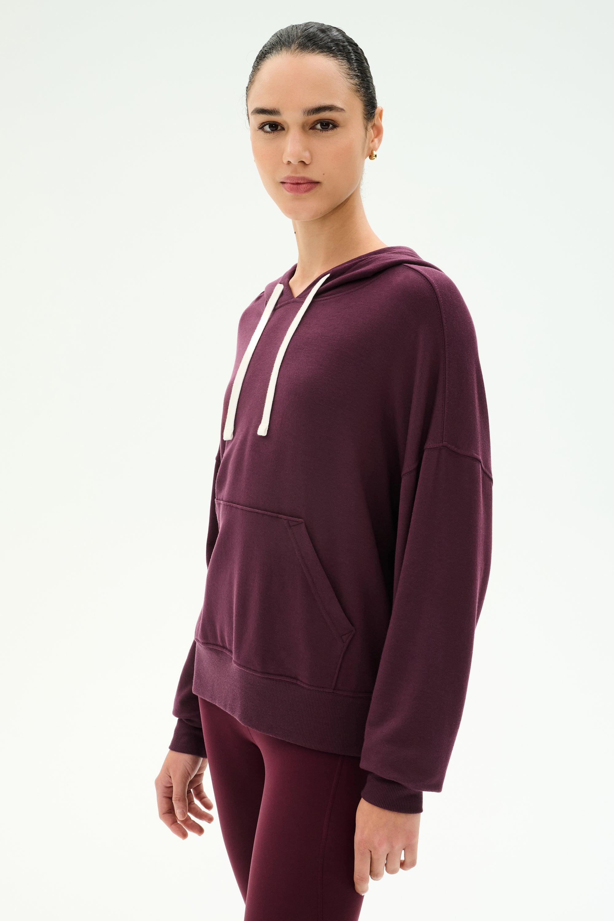 A woman wearing the SPLITS59 Joey Fleece Hoodie in Dark Cherry, featuring white drawstrings and matching leggings, crafted from a soft spandex blend, stands against a plain light background.