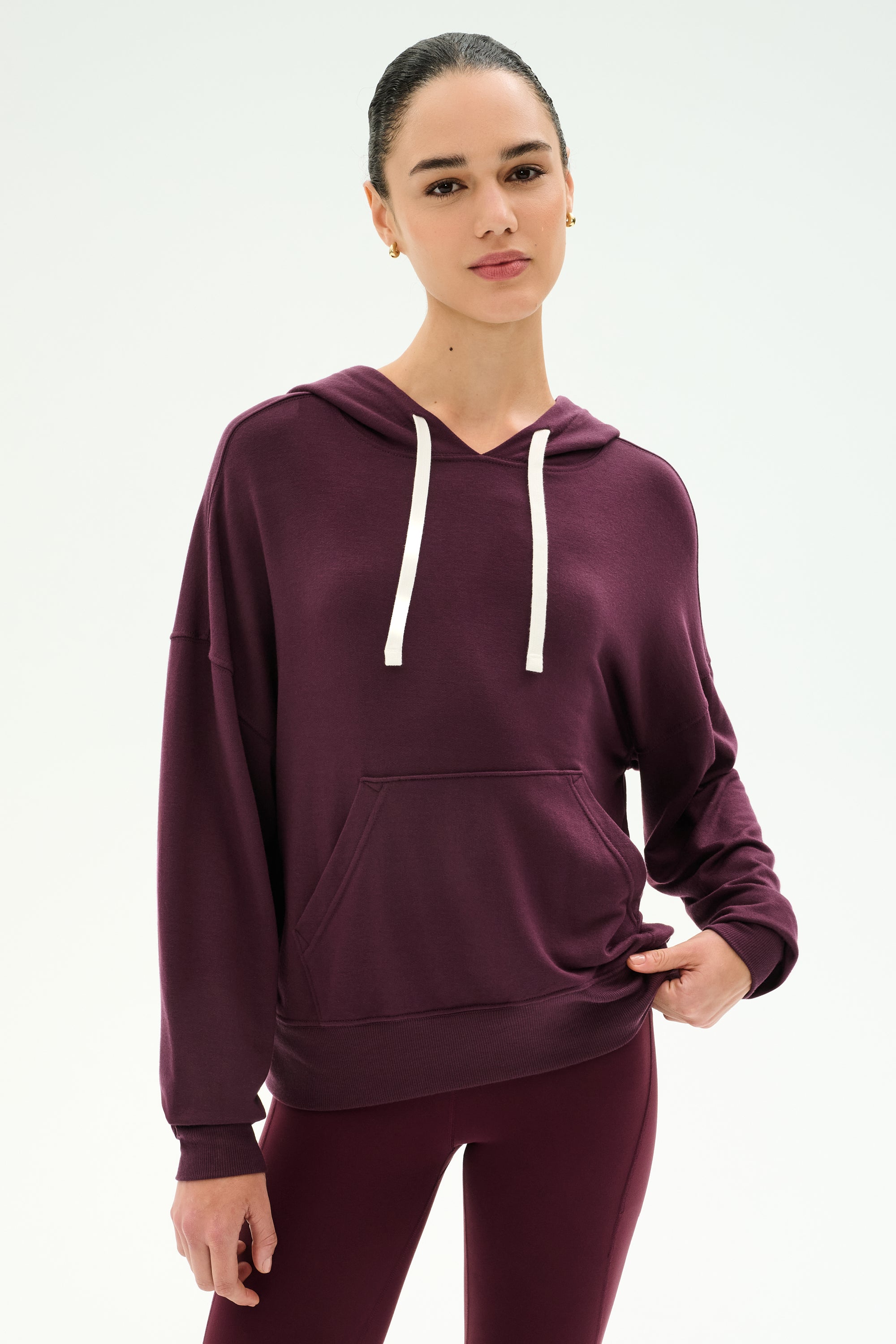 A woman wearing the SPLITS59 Joey Fleece Hoodie in Dark Cherry, featuring white drawstrings and paired with matching leggings, crafted from a soft modal fabric blend, stands against a plain white background.
