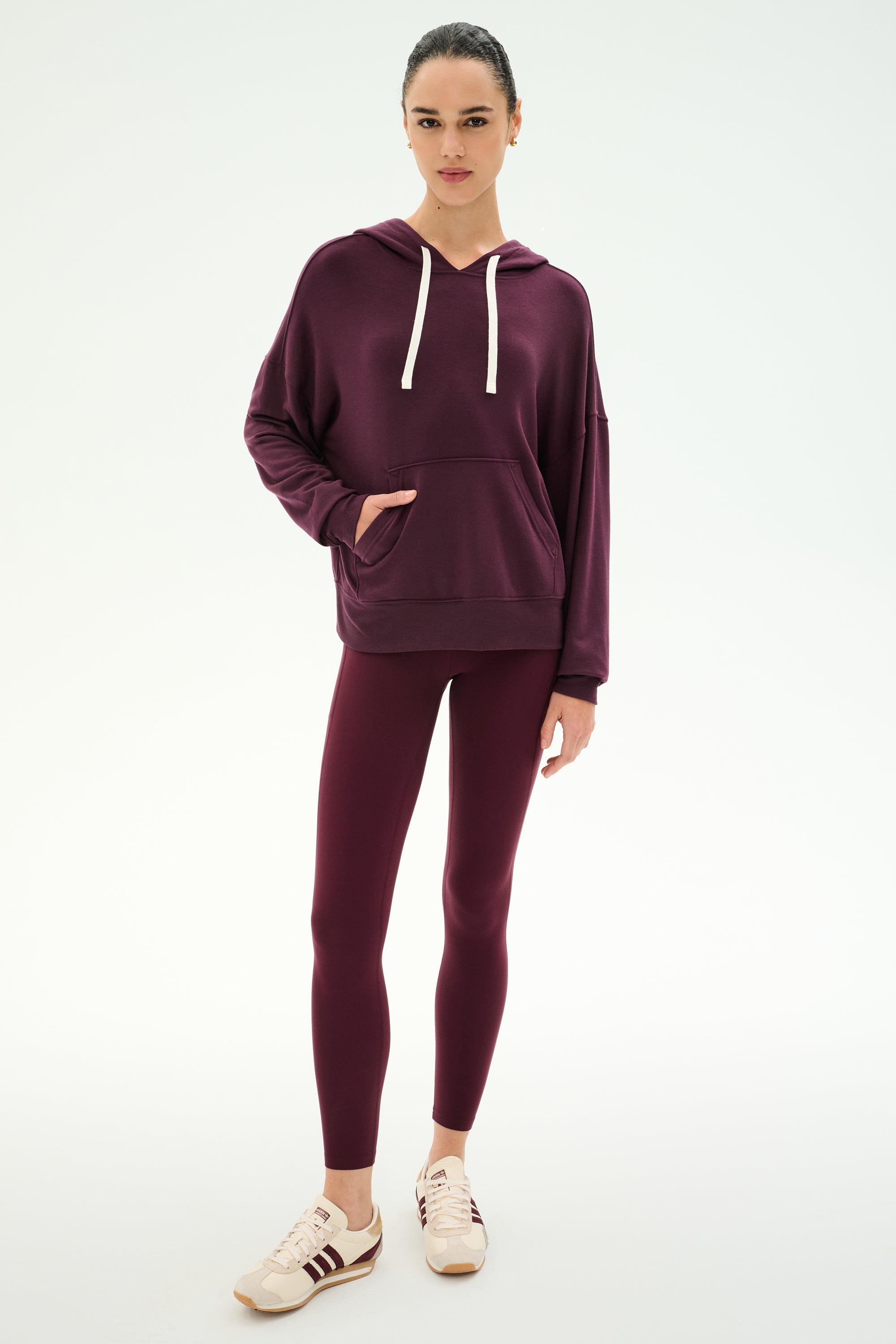 A woman stands against a plain white background wearing the SPLITS59 Joey Fleece Hoodie in Dark Cherry, featuring white drawstrings, paired with matching burgundy leggings crafted from a soft modal and spandex blend, and beige sneakers.