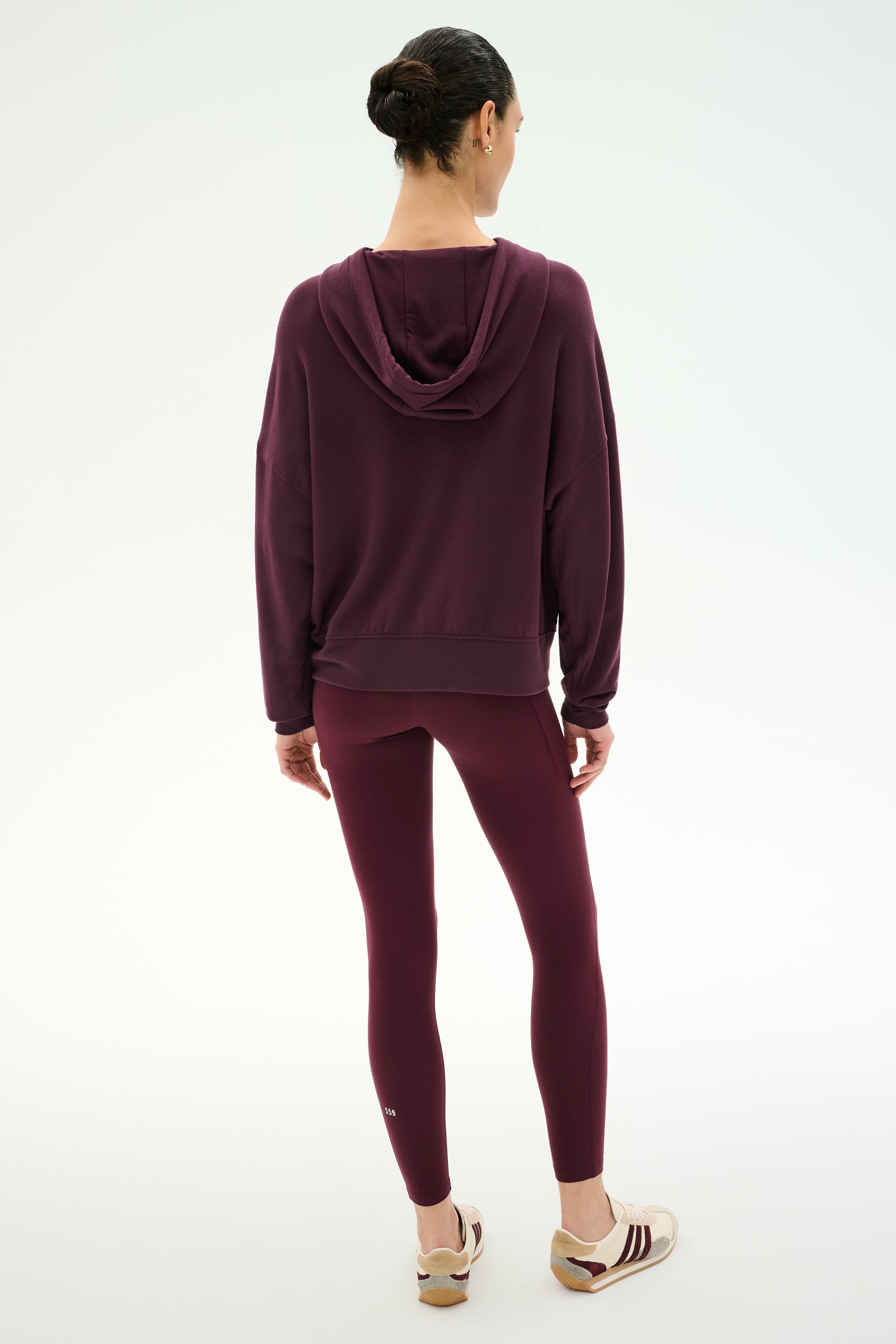 A person is seen from behind, standing against a plain background and wearing the SPLITS59 Joey Fleece Hoodie in Dark Cherry along with matching maroon leggings made from a comfortable spandex blend. Their outfit is completed with beige sneakers accented by maroon stripes.