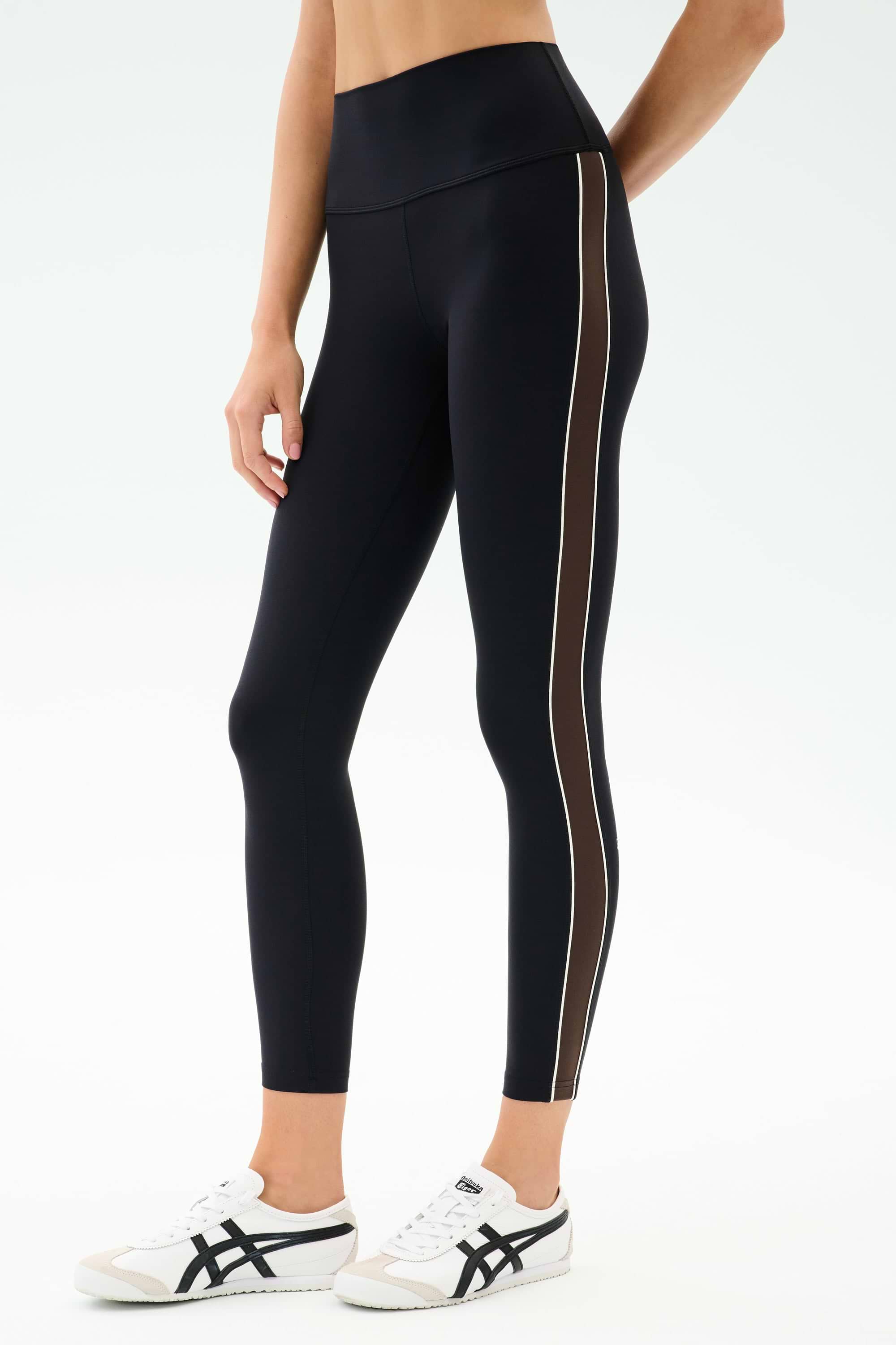 A person wears the SPLITS59 Margot Rigor Legging in Black/Dark Chocolate with brown and white side stripes, paired with white athletic shoes, standing against a plain background.