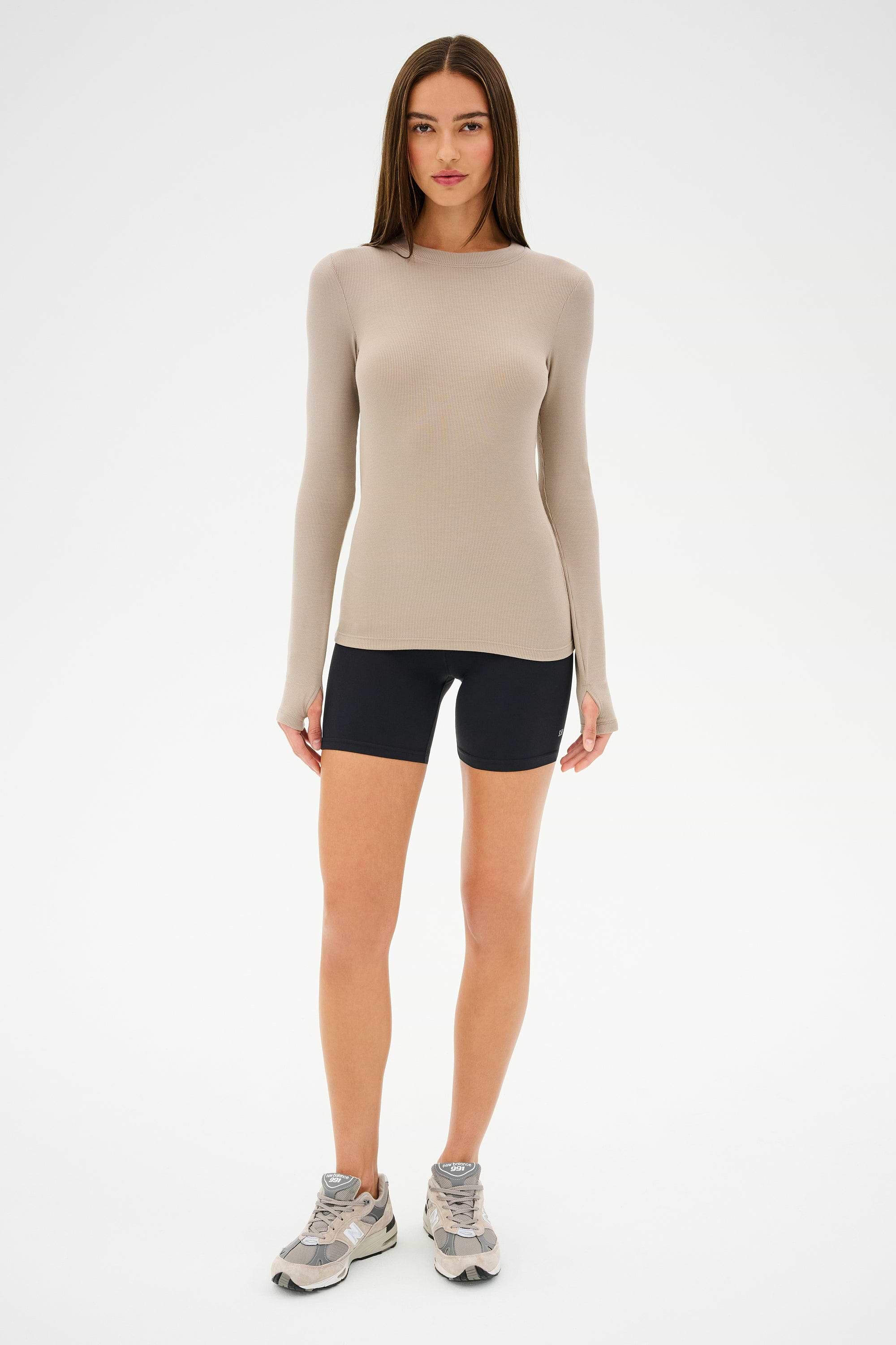 A person in a beige long-sleeve top, SPLITS59 Airweight High Waist Short in black, and gray sneakers stands against a white background.
