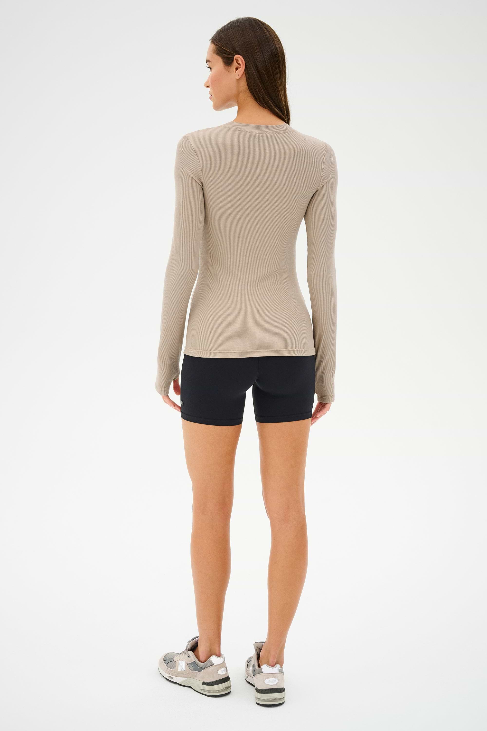 A person with long hair faces away in a beige long-sleeve top, wearing SPLITS59's Airweight High Waist Short in black and sneakers, set against a plain white background.