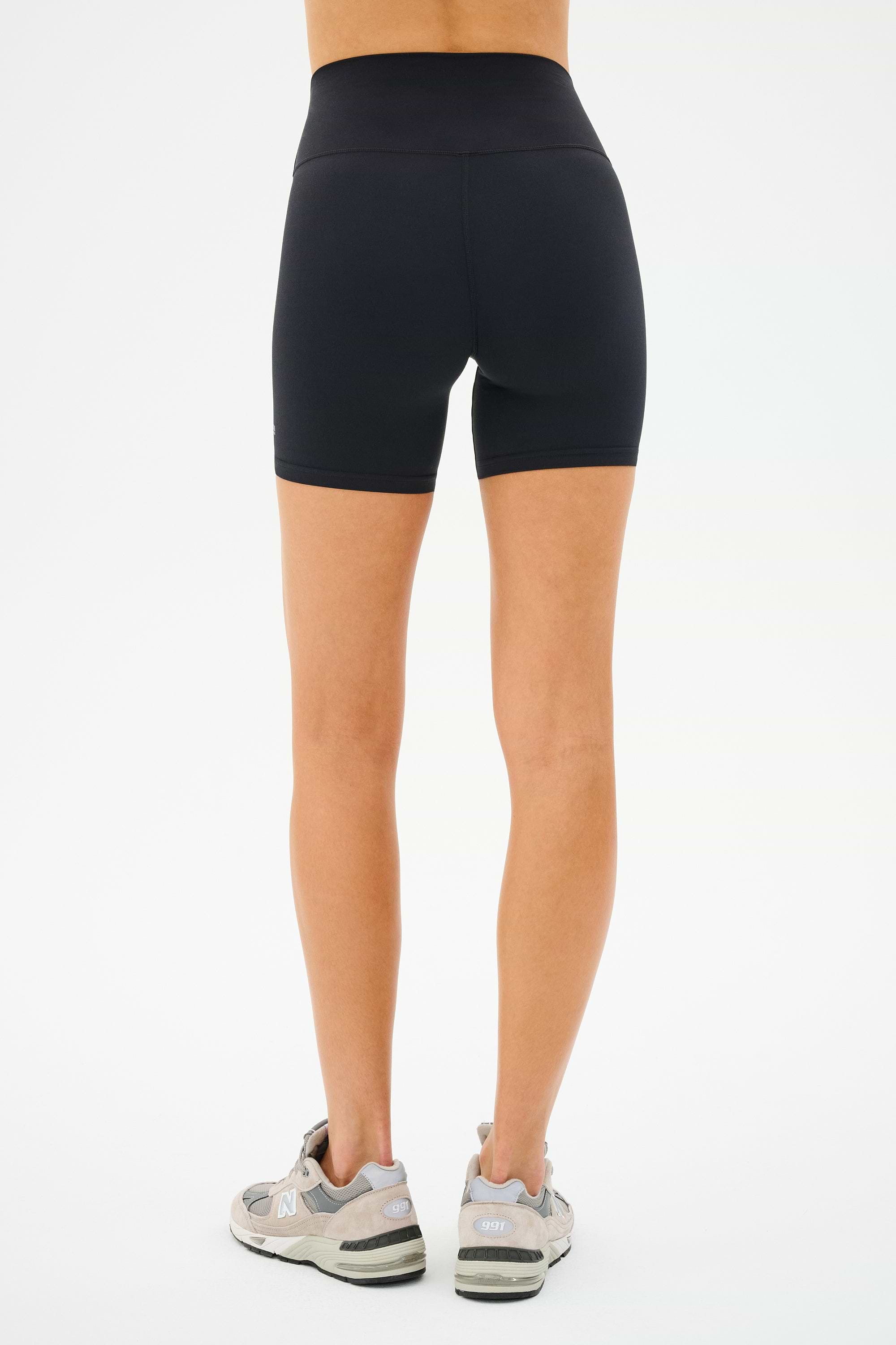 Viewed from the back, a person wears SPLITS59's Airweight High Waist Short in black and gray sneakers, ready for cycling against a plain white backdrop.
