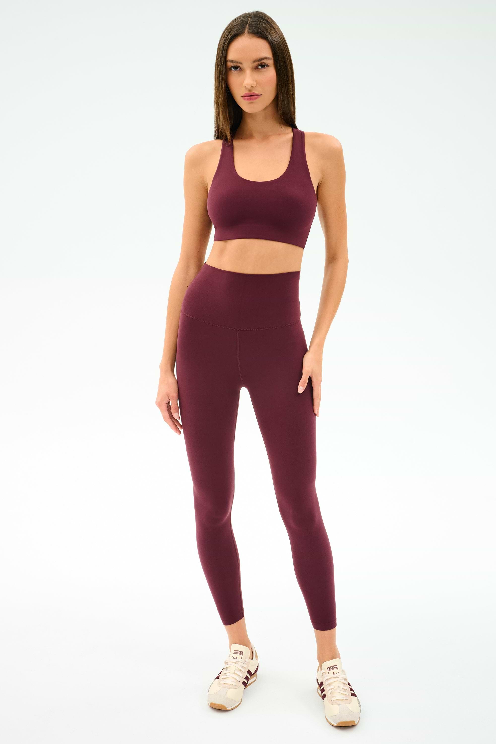 A woman stands against a plain background wearing the SPLITS59 Sara Airweight Bra in Dark Cherry paired with matching high-waisted leggings and light-colored sneakers.