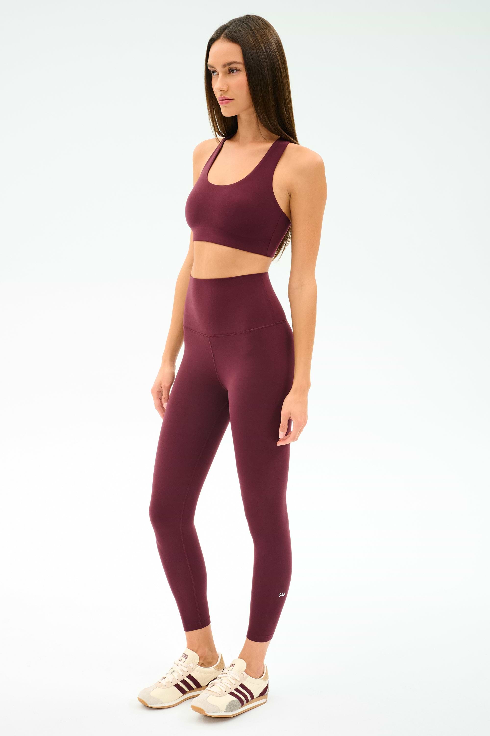 A woman wears the SPLITS59 Sara Airweight Bra in Dark Cherry with high-waisted spandex leggings and beige sneakers, standing against a plain light background.