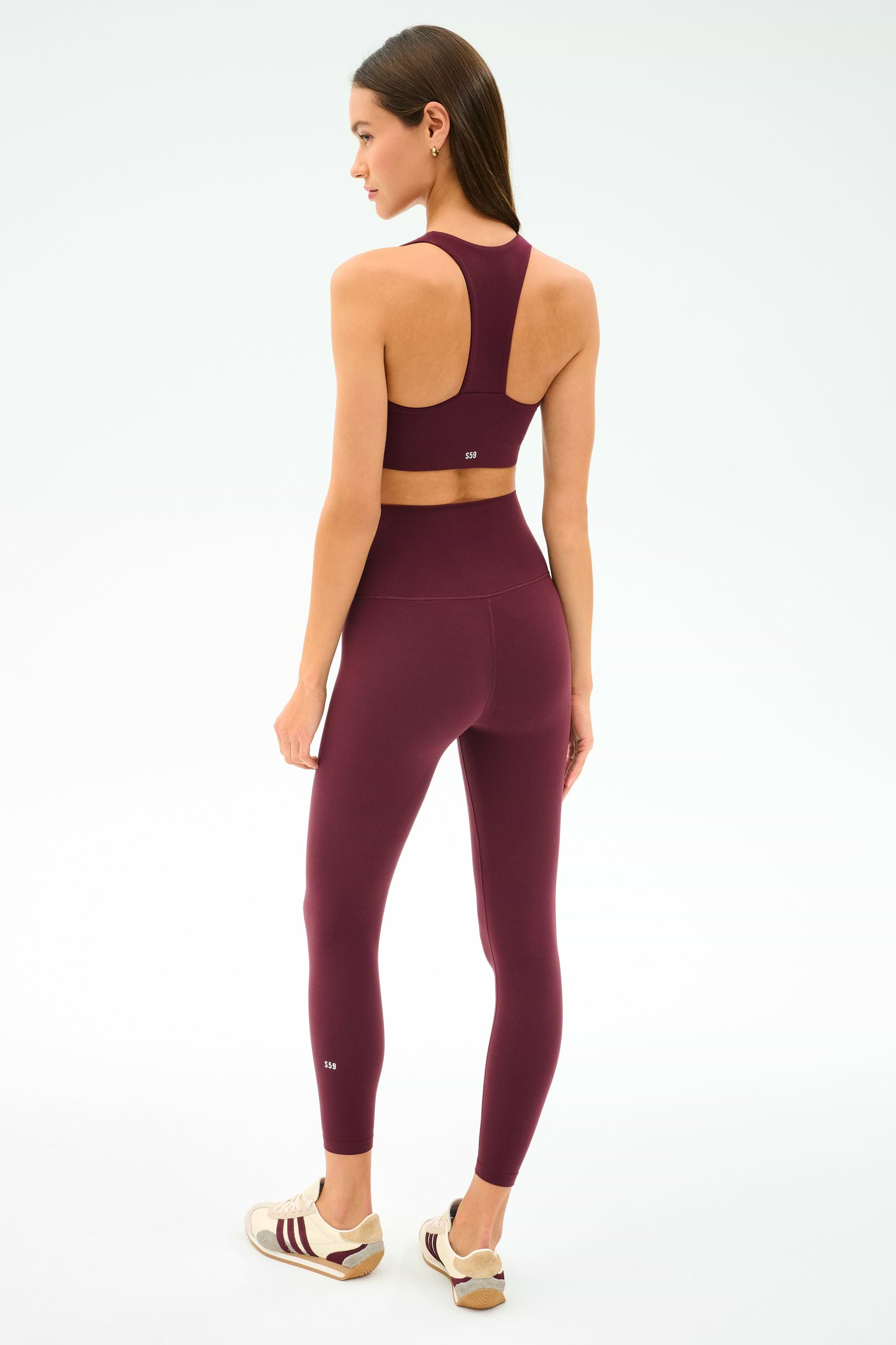 A woman faces away, wearing the SPLITS59 Sara Airweight Bra in Dark Cherry with matching leggings and white sneakers, standing against a plain light background.