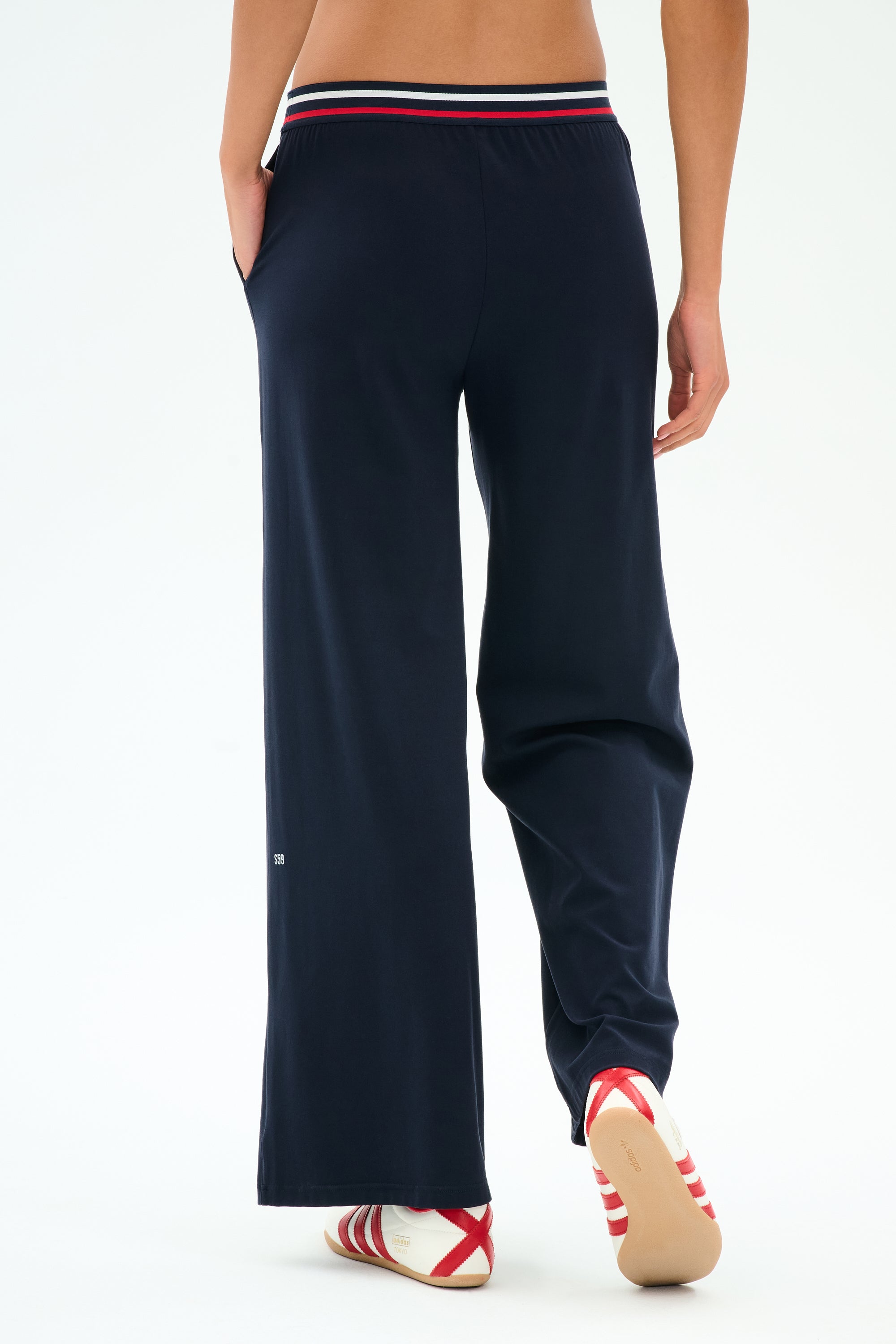 A person is shown from the back wearing SPLITS59 Michele Airweight Trouser in Indigo with hands in pockets and white slip-on shoes featuring red and navy stripes, standing on a white background.