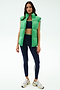 A woman wears the SPLITS59 Summit Nylon Puffer Vest in Grass over a navy sports bra, striped navy leggings, and white sneakers—ideal for cool weather workouts—shown against a plain light background.