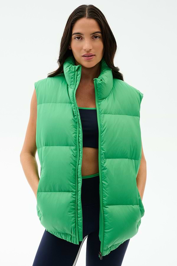 Wearing the SPLITS59 Summit Nylon Puffer Vest in Grass over a dark sports bra and leggings, this person stands against a plain light background—ready for cool weather workouts with lightweight warmth and style.