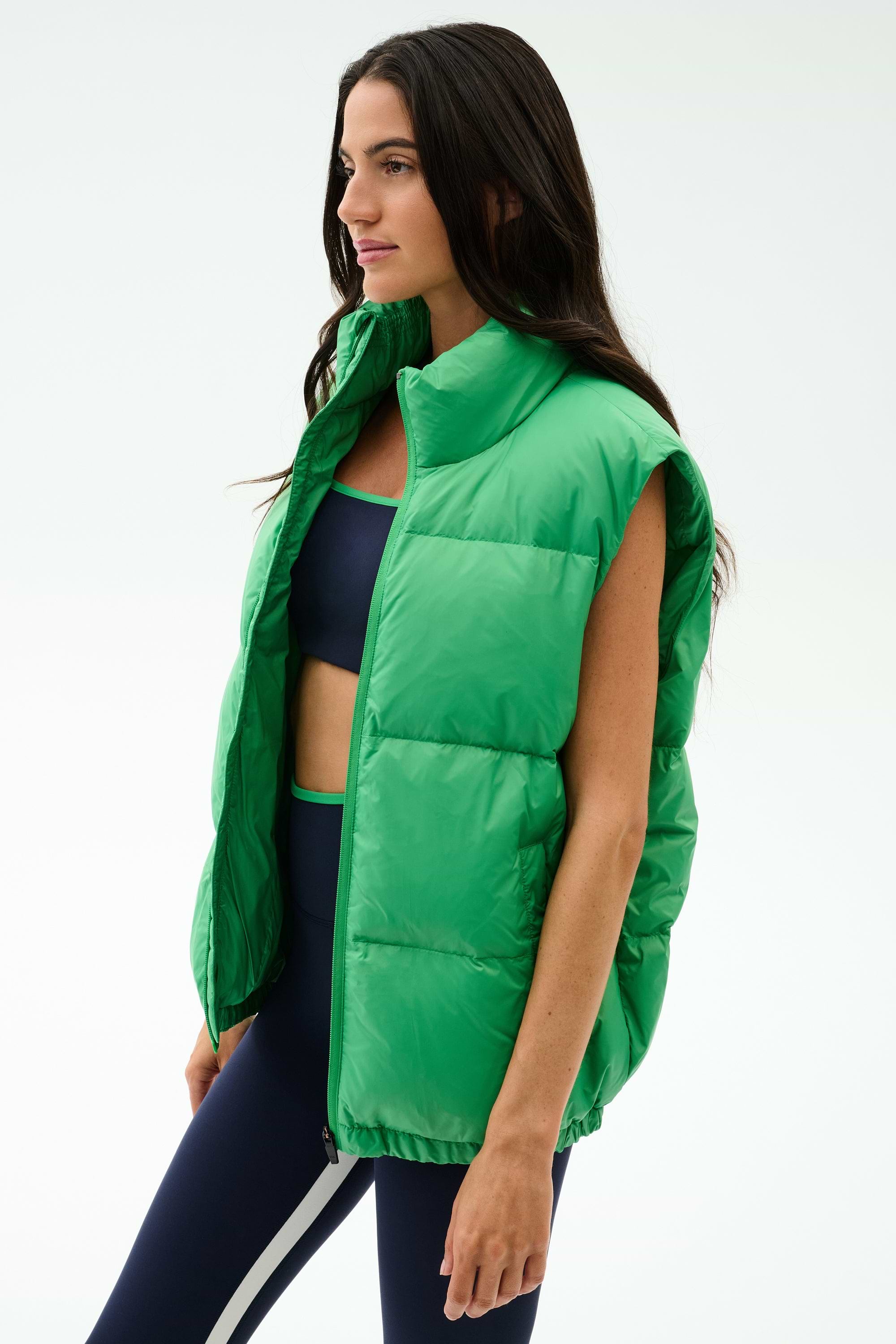 A woman wears the SPLITS59 Summit Nylon Puffer Vest in Grass over a navy sports bra and navy leggings with a white stripe, standing against a plain white background—ideal for cool weather workouts with its lightweight insulation.