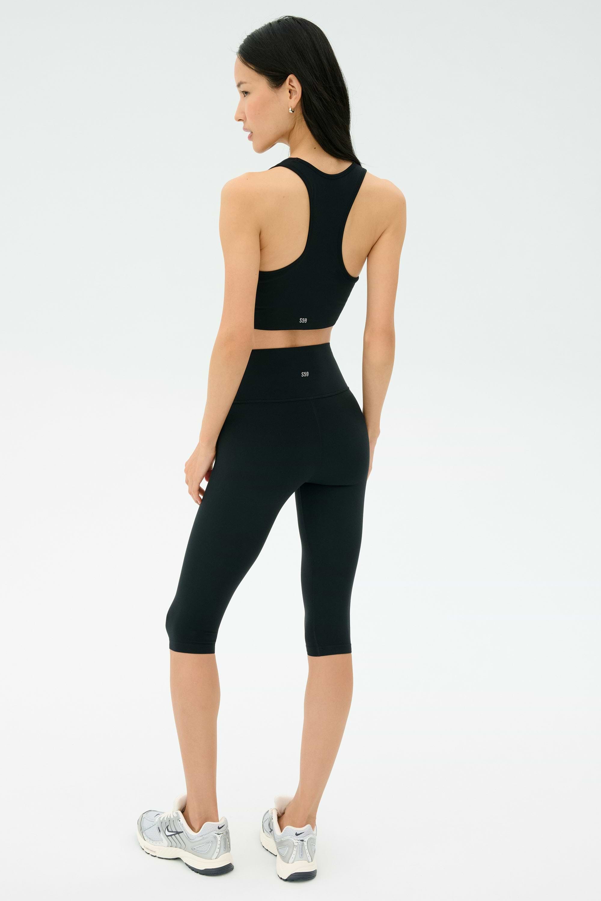 A woman stands on a white background, shown from the back, wearing a black sports bra and SPLITS59 Airweight High Waist 16" Legging in black with silver athletic shoes.