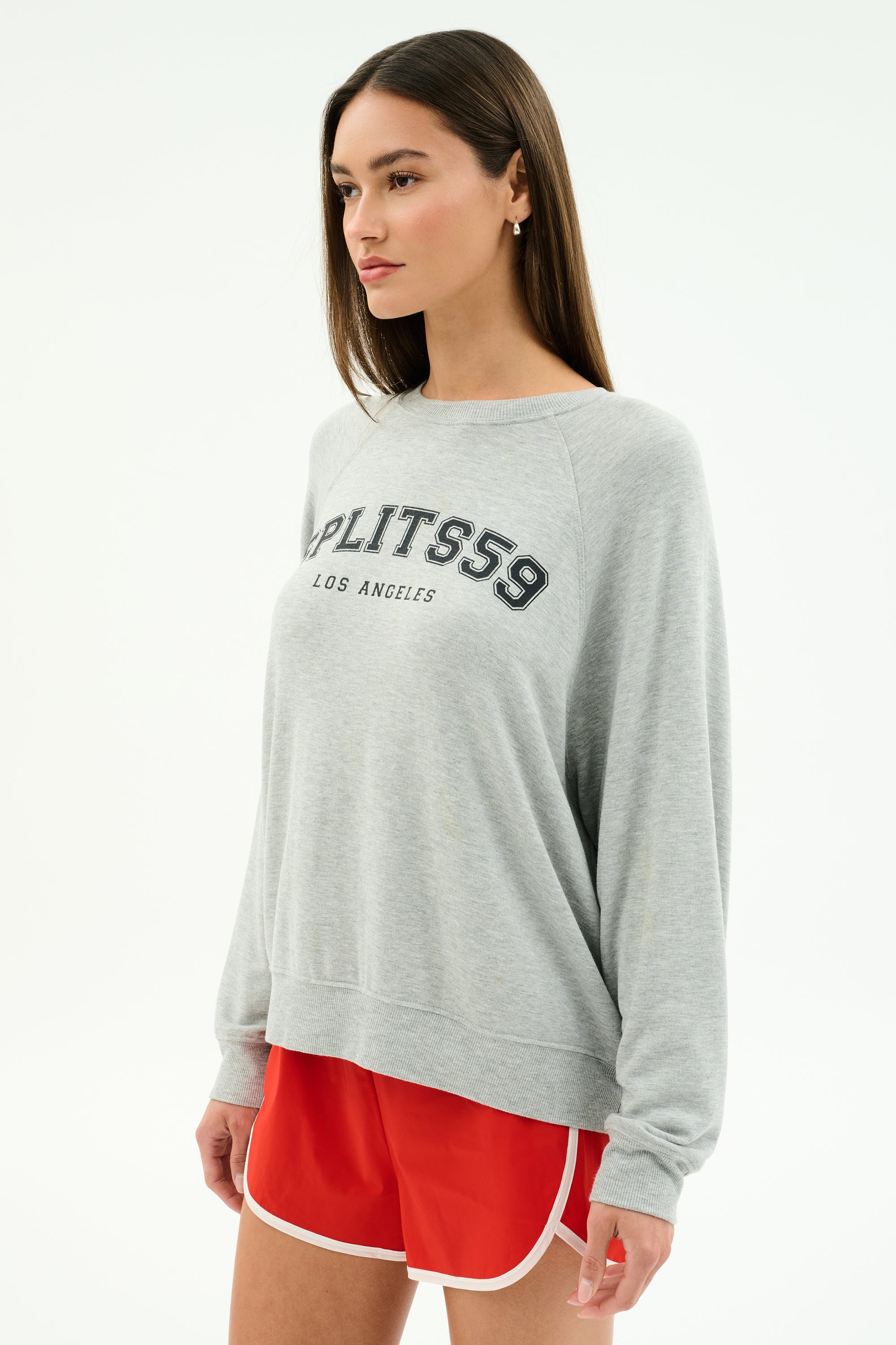 A woman stands against a white background wearing the SPLITS59 Andie Oversized Fleece Varsity Graphic Sweatshirt in heather grey with "Splits59 Los Angeles" text and red athletic shorts.