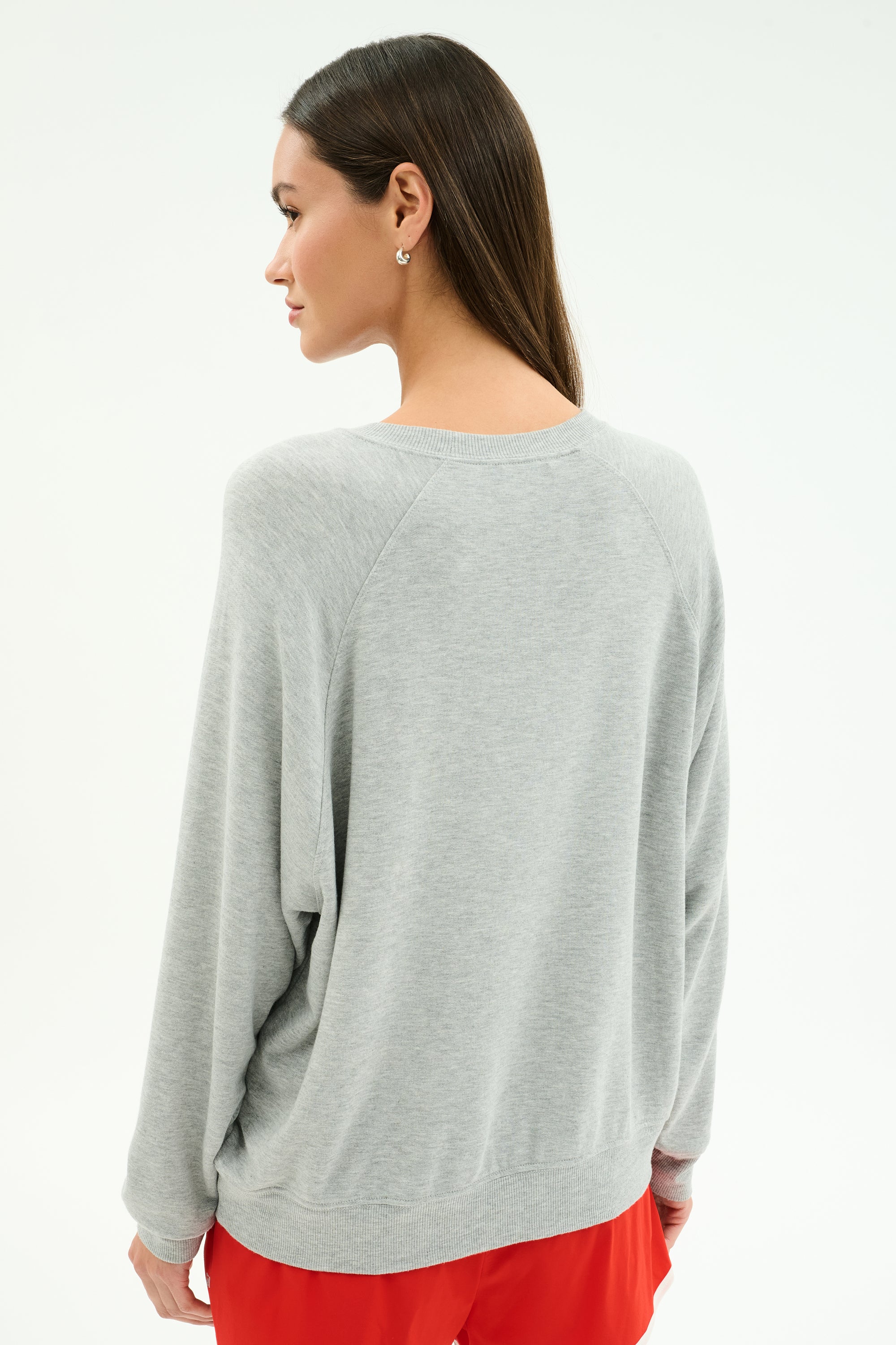 A woman stands with her back to the camera wearing the SPLITS59 Andie Oversized Fleece Varsity Graphic Sweatshirt in Heather Grey, paired with red shorts, set against a plain light background.