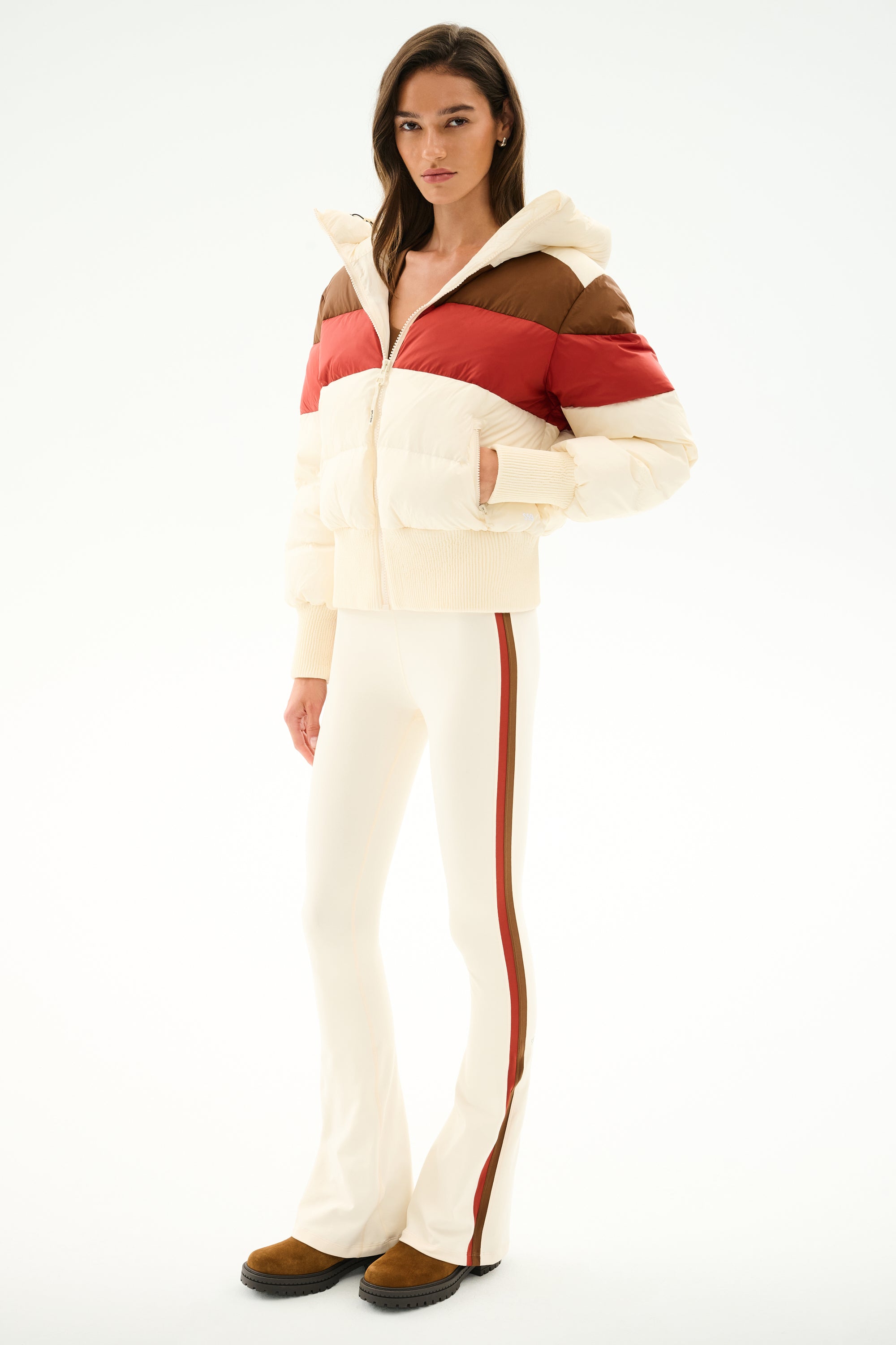 Woman wearing the Aspen Puffer Jacket - Creme/Terracotta by SPLITS59, cream flared pants with red side stripe, and brown shoes against a white background.