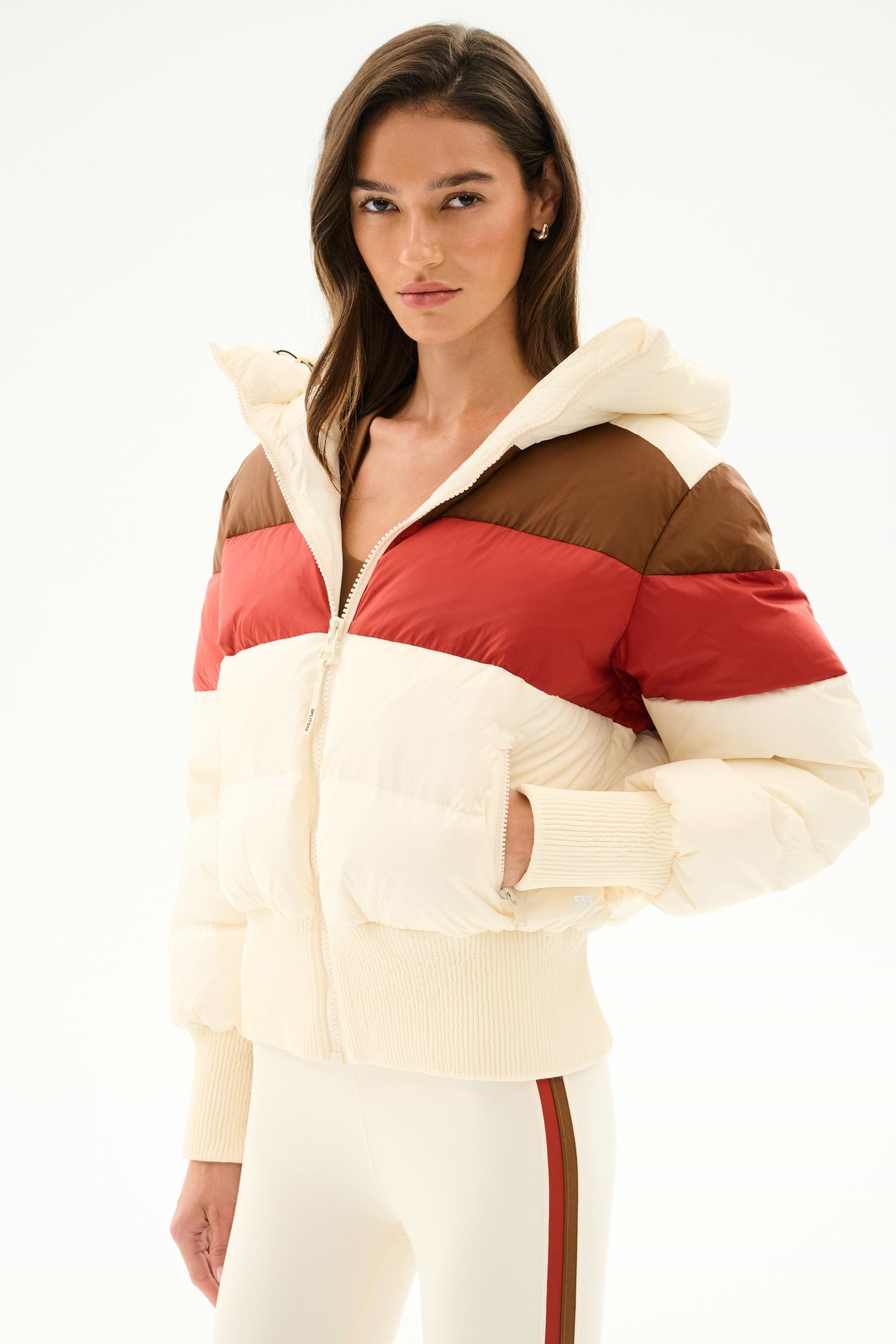 Model wears the SPLITS59 Aspen Puffer Jacket - Creme/Terracotta, featuring a nylon body with white duck down fill, paired with striped cream pants against a white background.