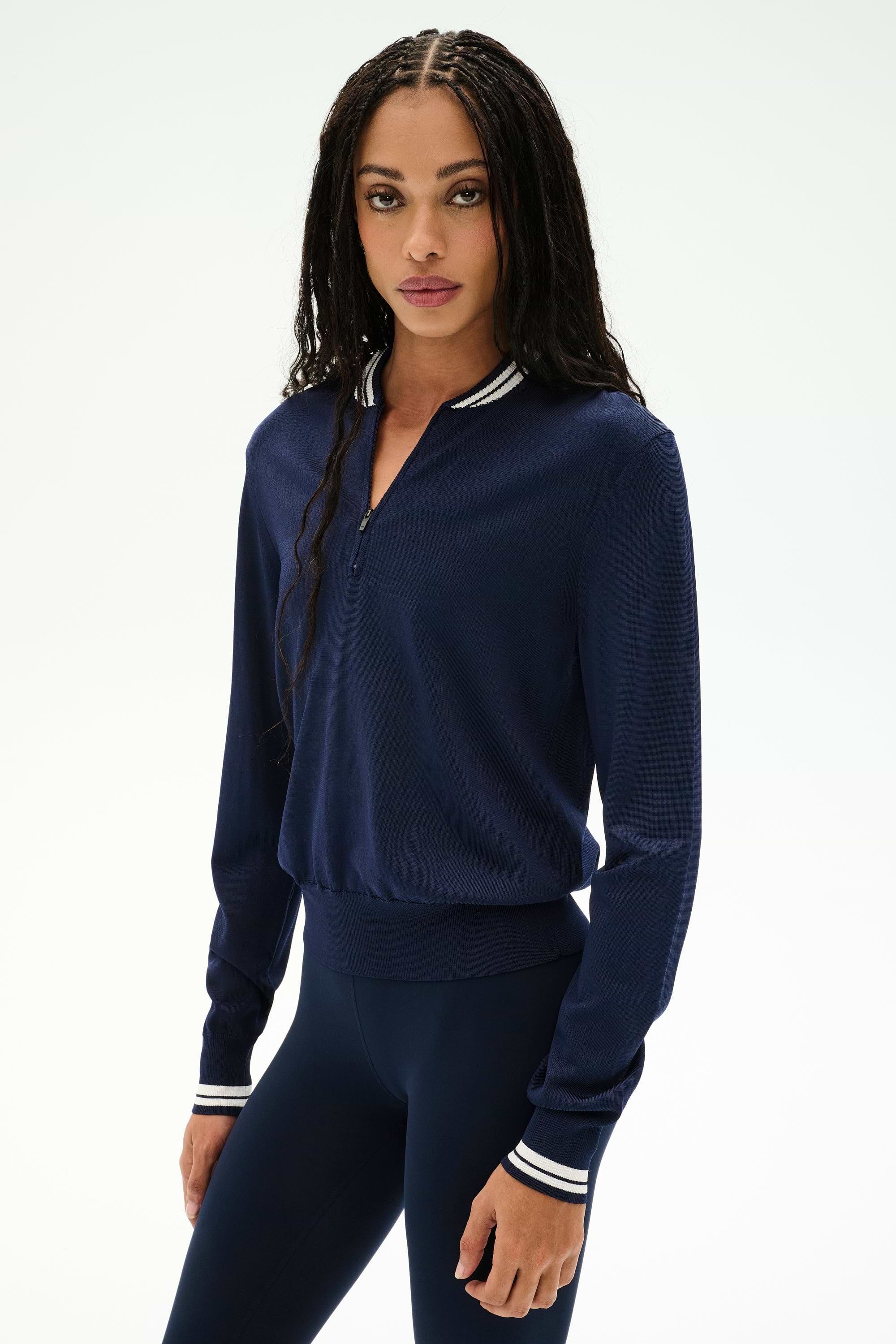 A woman with long braided hair wears the SPLITS59 Bonnie Knit Half Zip in Indigo/White, paired with matching navy leggings, both crafted from a soft rayon-nylon blend, as she stands against a plain light background.