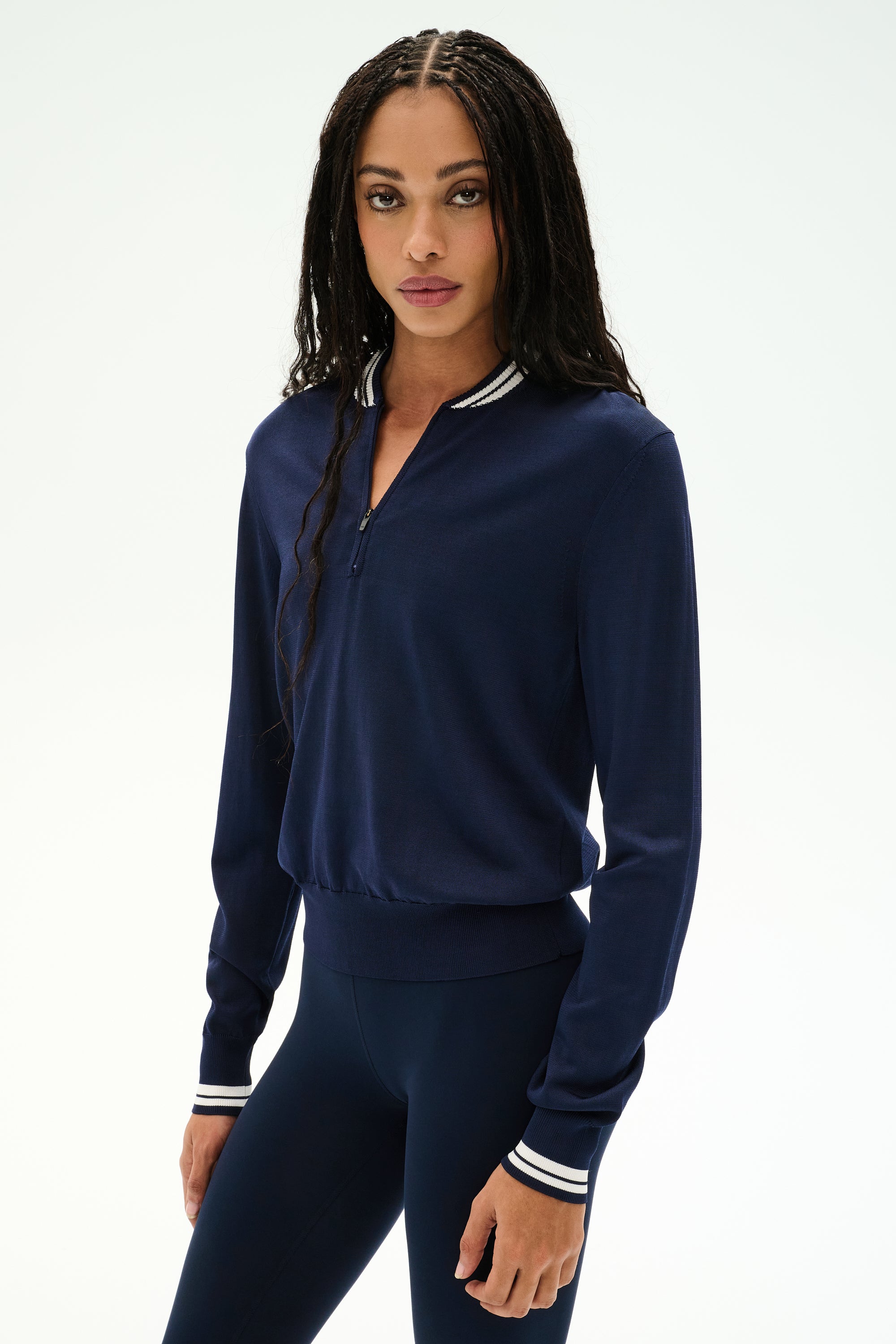 A woman with long braided hair wears the SPLITS59 Bonnie Knit Half Zip in Indigo/White, paired with matching navy leggings, both crafted from a soft rayon-nylon blend, as she stands against a plain light background.