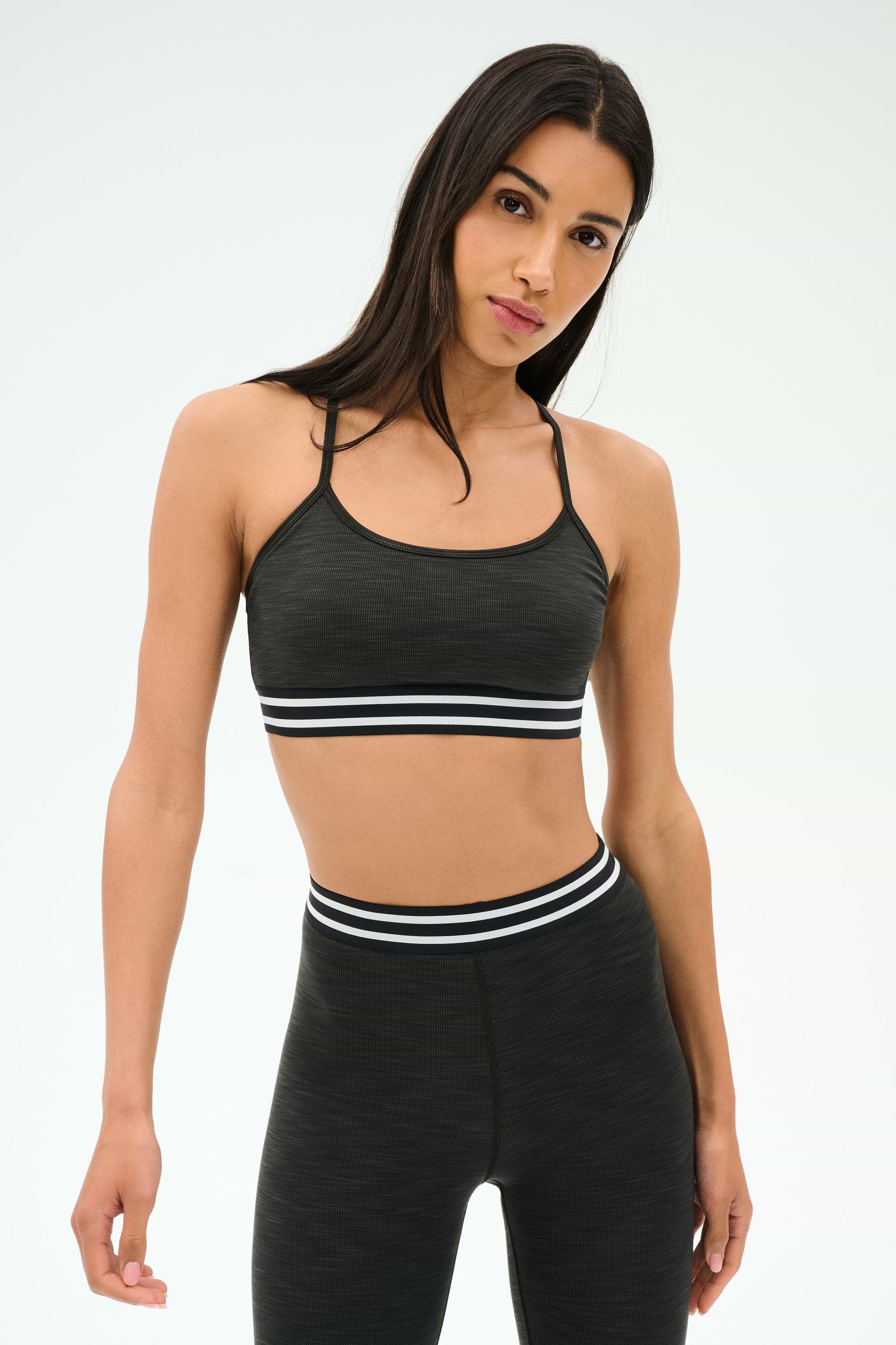 A person models the SPLITS59 Cali Active Rib Bra in Heather Graphite, featuring a supportive dark design, against a plain light background.