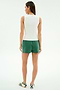 A woman faces away, wearing SPLITS59's Ella Airweight Track Short in Arugula/White with a sleeveless white top and white sneakers with green stripes, set against a plain light background.