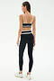 A woman stands facing away, ready to work out in SPLITS59's Mason High Waist Rigor Full Length leggings in Black/White and a black sports bra with white straps, paired with white sneakers against a plain background.
