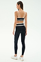 A woman stands facing away, ready to work out in SPLITS59's Mason High Waist Rigor Full Length leggings in Black/White and a black sports bra with white straps, paired with white sneakers against a plain background.