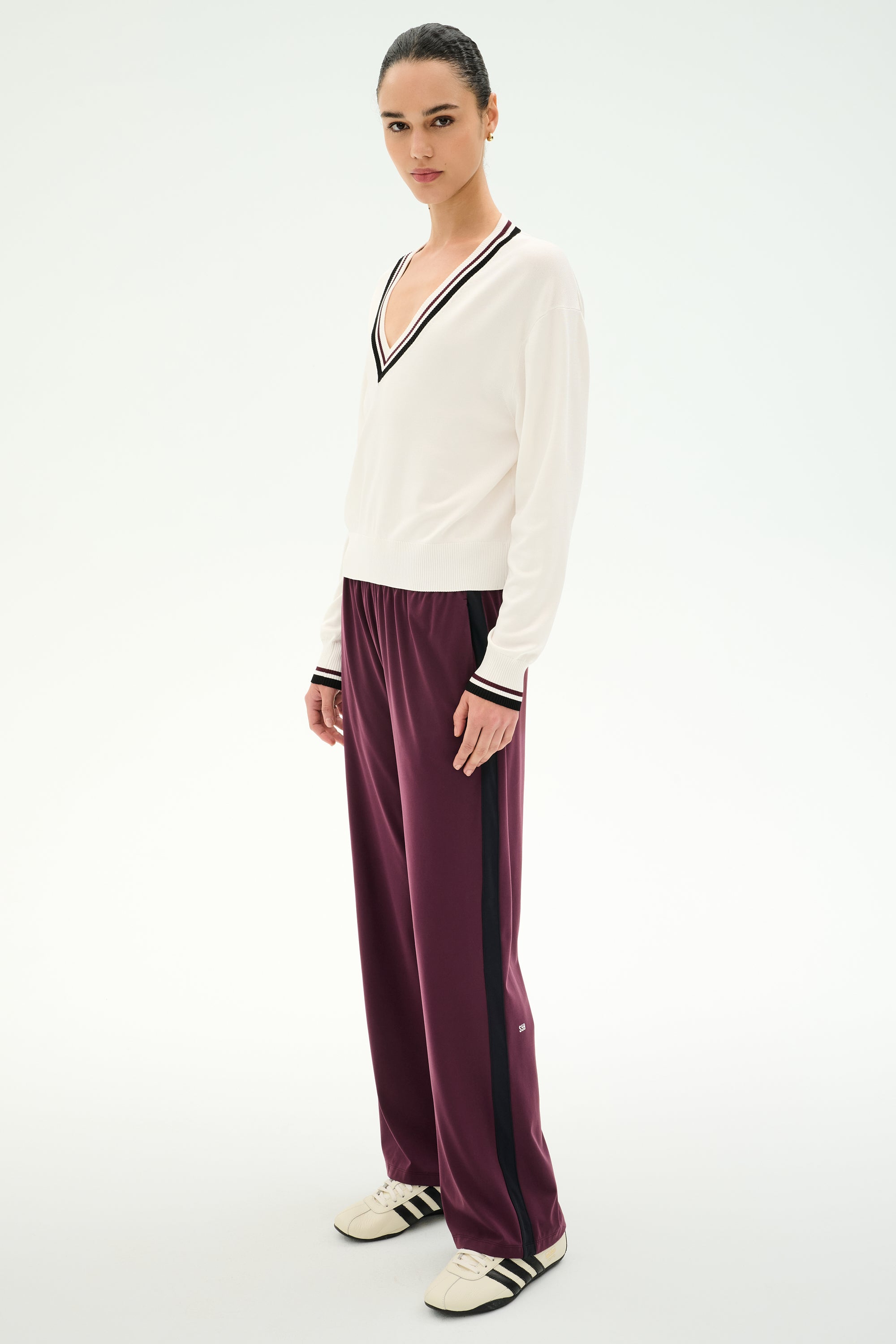 A person is seen standing on a plain white background, wearing the SPLITS59 Jemma Knit V Neck in White/Black—an ultra-soft nylon-rayon blend sweater with black trim—styled with maroon track pants featuring black stripes and white sneakers.