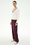 Person standing against a plain background, wearing a white V-neck sweater with dark trim, SPLITS59 Maxie Rigor Track Pant in Dark Cherry/Black, and beige Adidas sneakers with black stripes.