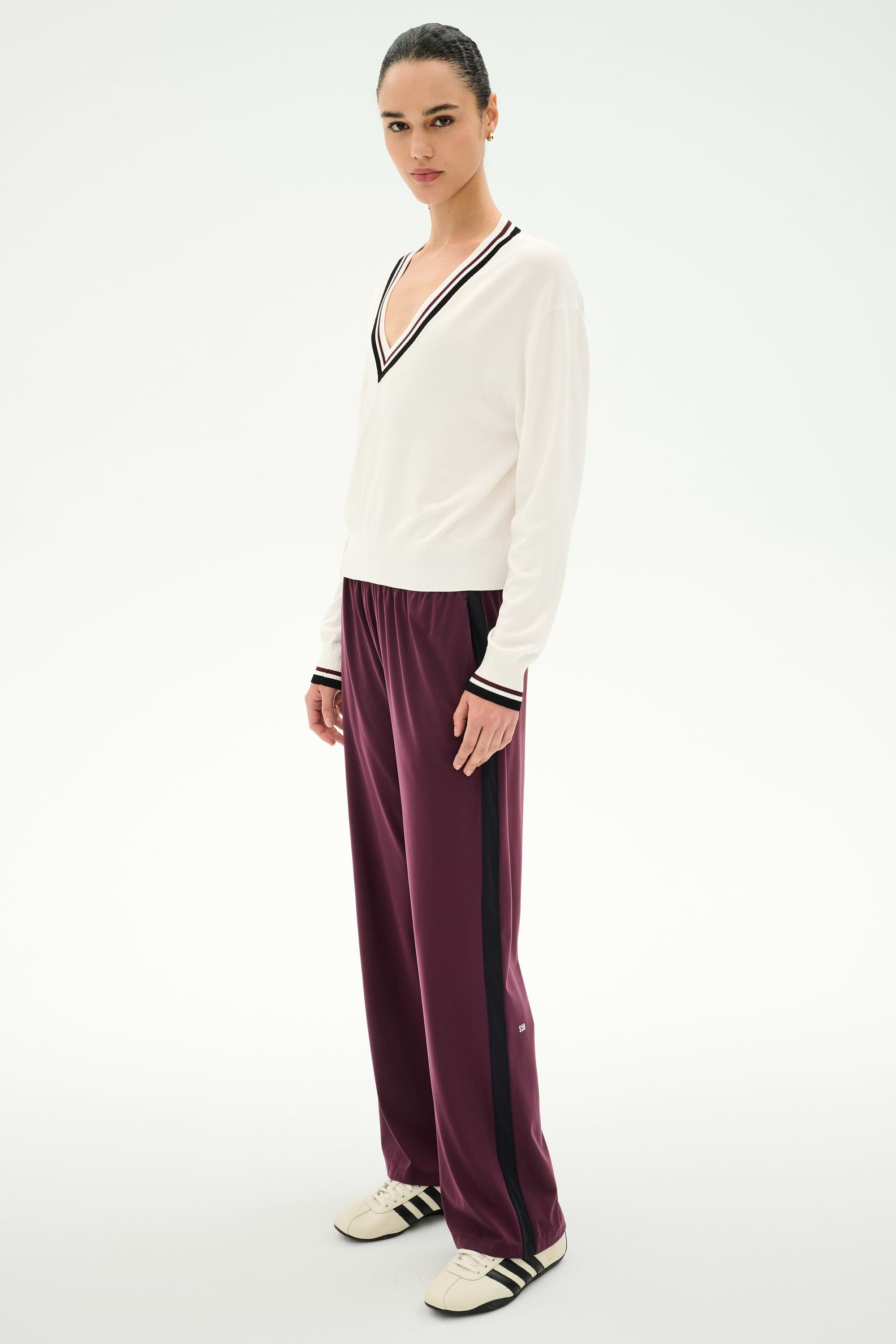 Person standing against a plain background, wearing a white V-neck sweater with dark trim, SPLITS59 Maxie Rigor Track Pant in Dark Cherry/Black, and beige Adidas sneakers with black stripes.