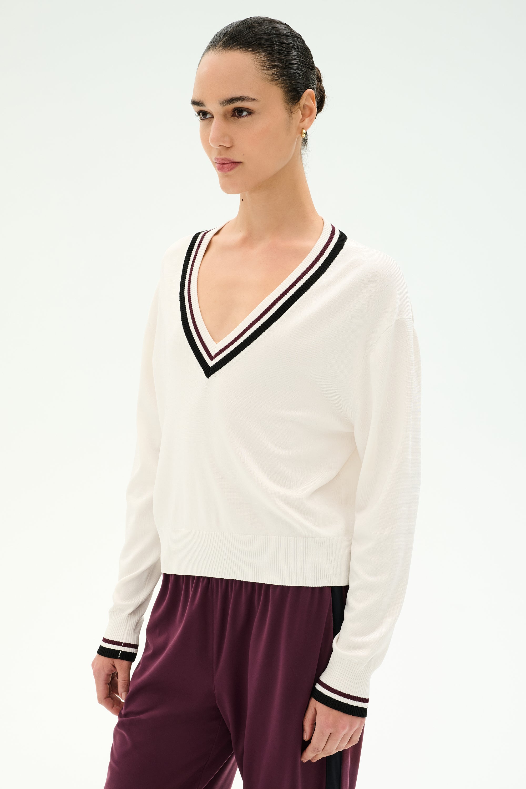 A woman stands against a plain light background wearing the SPLITS59 Jemma Knit V Neck in White/Black—a soft rayon-nylon blend sweater featuring a white V-neck design with black trim—paired with maroon pants.