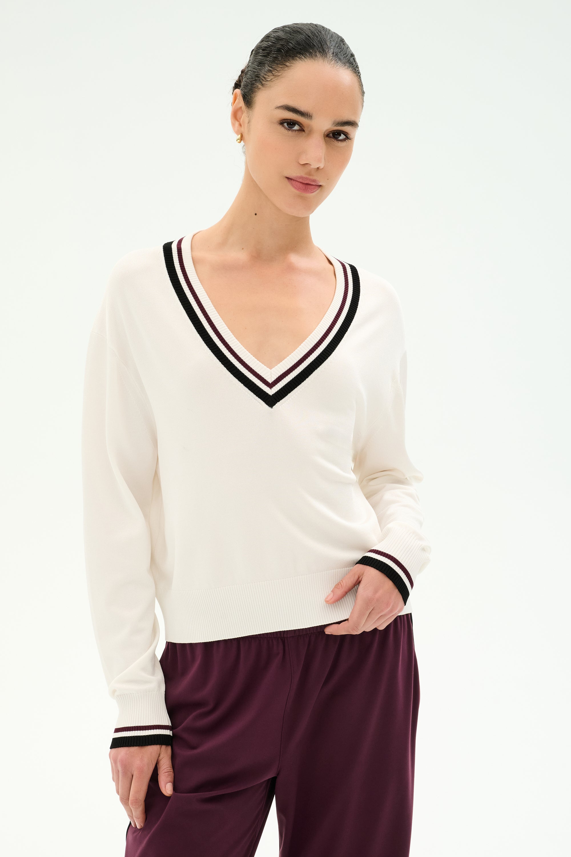 A woman is wearing the SPLITS59 Jemma Knit V Neck in White/Black—a soft, comfortable sweater crafted from a rayon and nylon blend—paired with maroon pants and standing against a plain light background.