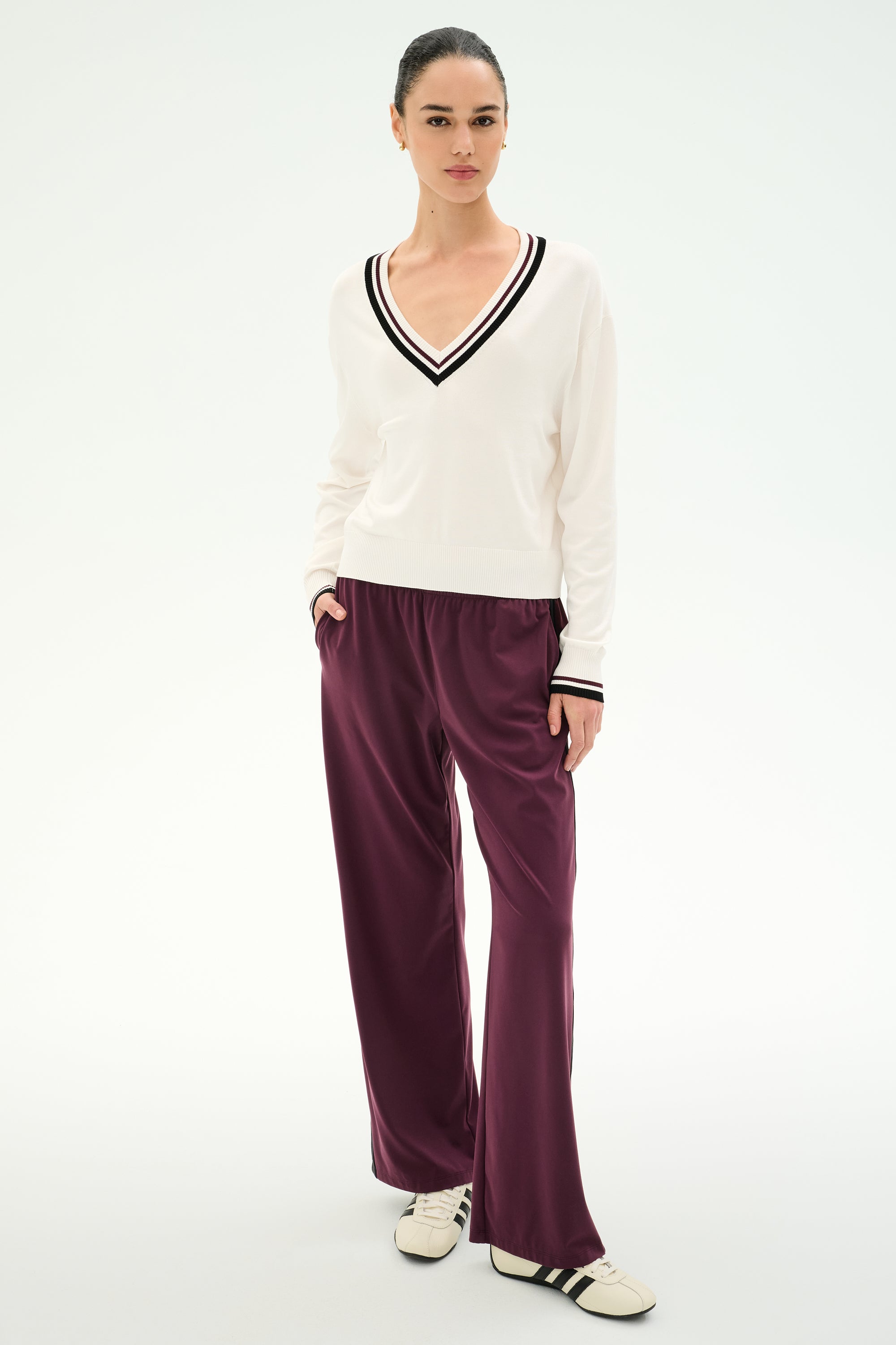 Wearing the SPLITS59 Jemma Knit V Neck in White/Black, this person pairs the sweater with loose maroon rayon-blend pants and white sneakers featuring black stripes, set against a plain light background.