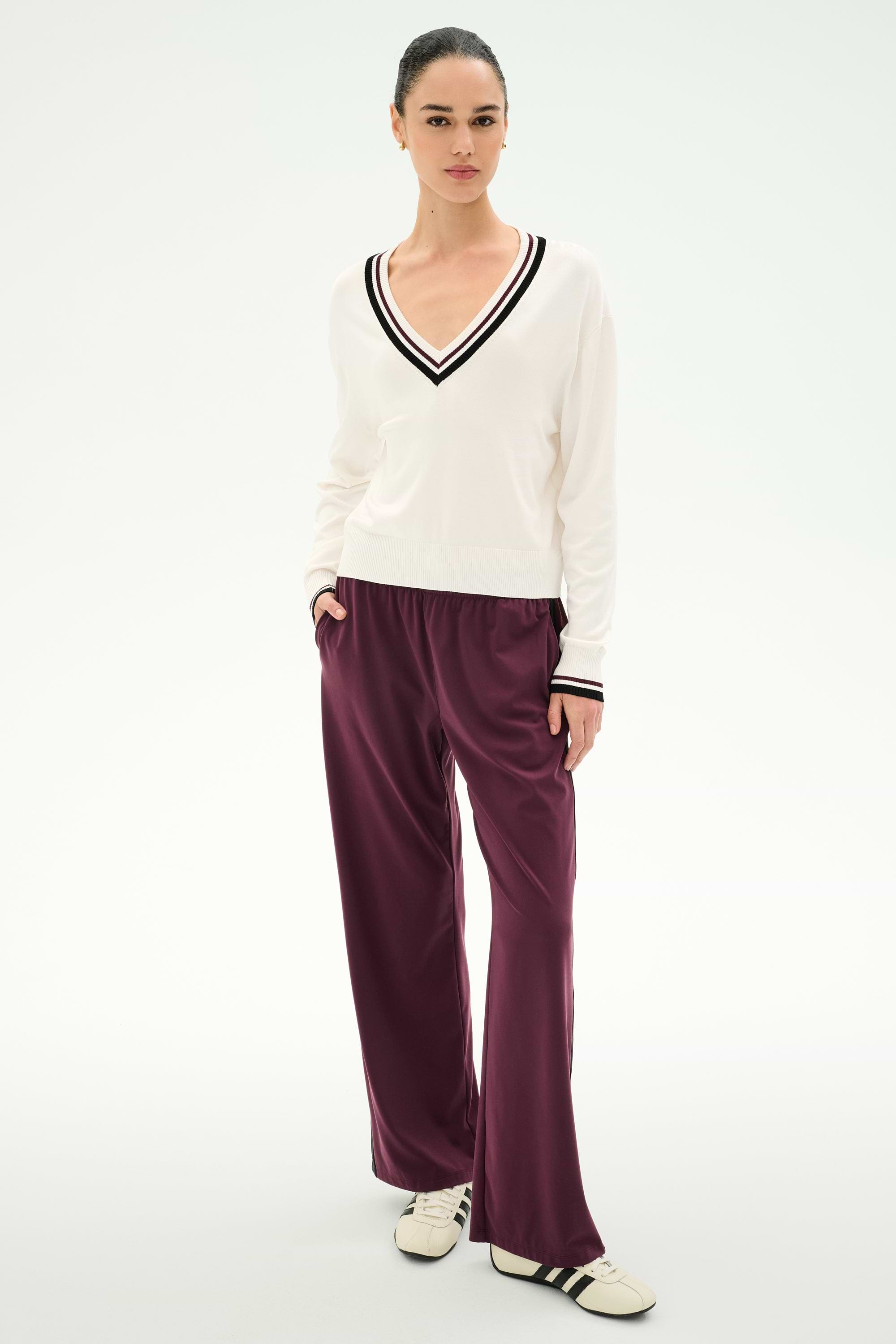 A woman stands against a white background, wearing a white V-neck sweater with black stripes, SPLITS59 Maxie Rigor Track Pant in Dark Cherry/Black, and white sneakers with black stripes. Her hands are in her pockets and her hair is tied back.