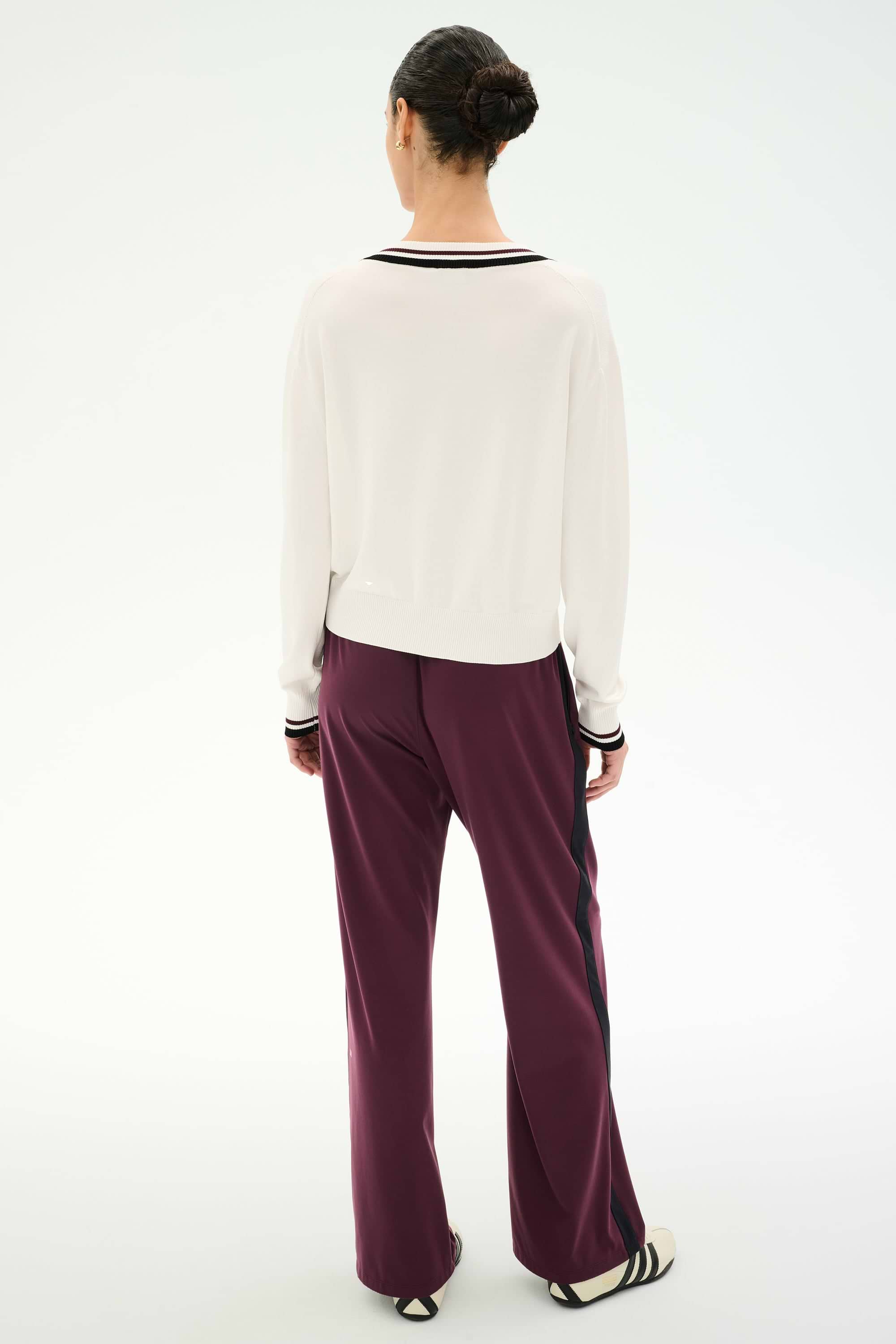 A person stands with their back to the camera, wearing a white long-sleeve spandex top, SPLITS59 Maxie Rigor Track Pants in Dark Cherry/Black, and flat shoes. Their hair is styled in a neat bun against a plain white background.