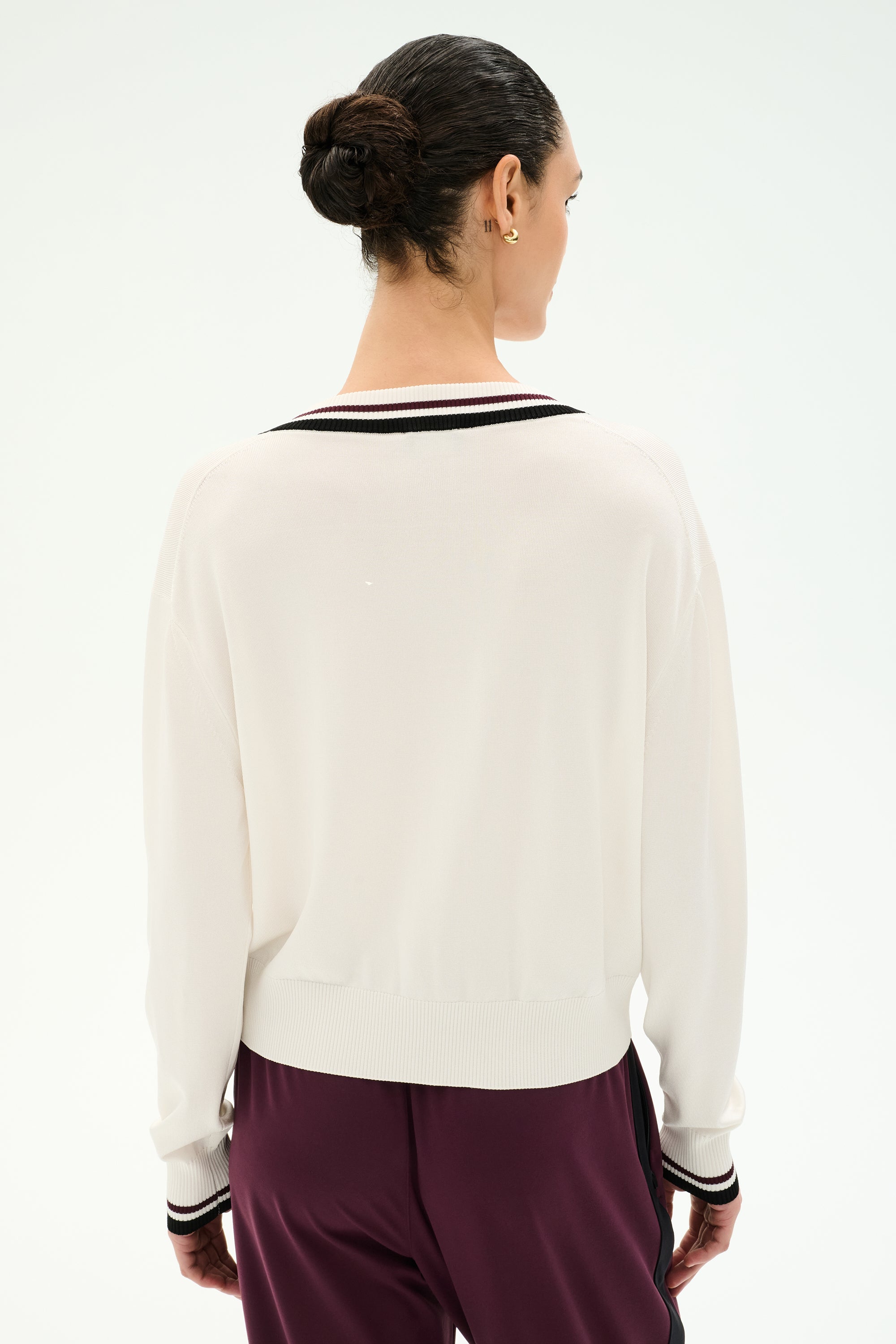 A person stands with their back to the camera, wearing the SPLITS59 Jemma Knit V Neck in white with black trim, crafted from a soft rayon blend, paired with burgundy pants against a plain light background.