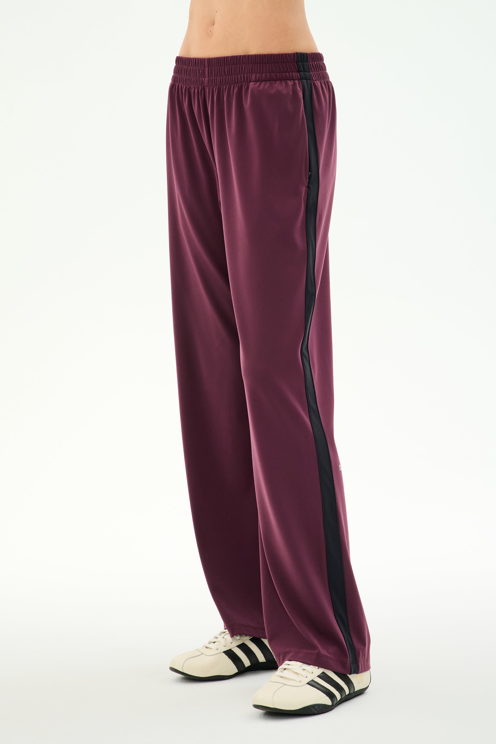 A person wearing the SPLITS59 Maxie Rigor Track Pant in Dark Cherry/Black, featuring a loose fit and a comfortable nylon-spandex blend with a black side stripe, paired with white sneakers with black stripes, stands against a plain white background.