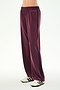 A person wearing the SPLITS59 Maxie Rigor Track Pant in Dark Cherry/Black, featuring a loose fit and a comfortable nylon-spandex blend with a black side stripe, paired with white sneakers with black stripes, stands against a plain white background.